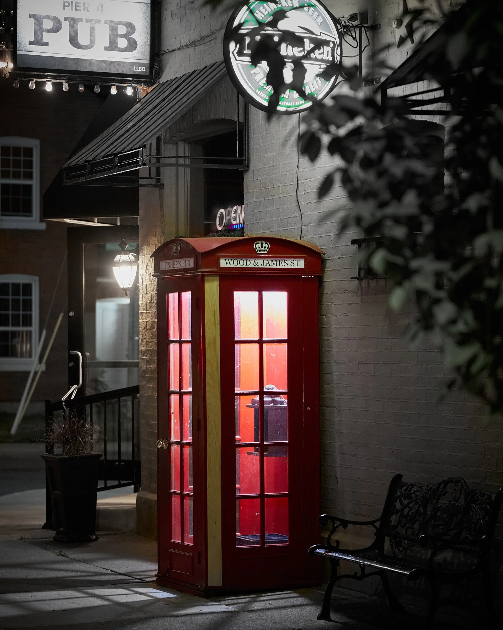 A red British-style telephone booth with interior lighting, located outside on a sidewalk, next to a black metal bench and potted plant, with signs for a pub and neon signs on a brick building at night.