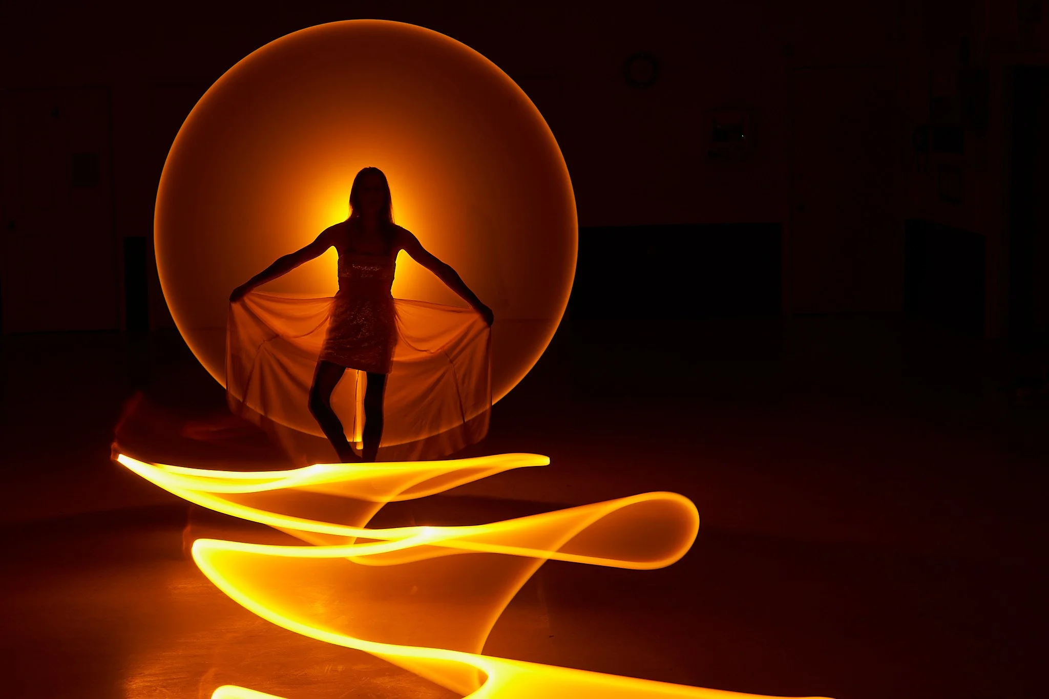 A silhouette of a woman in a dress standing in front of a glowing orange orb with light trails around her, creating a light painting effect.