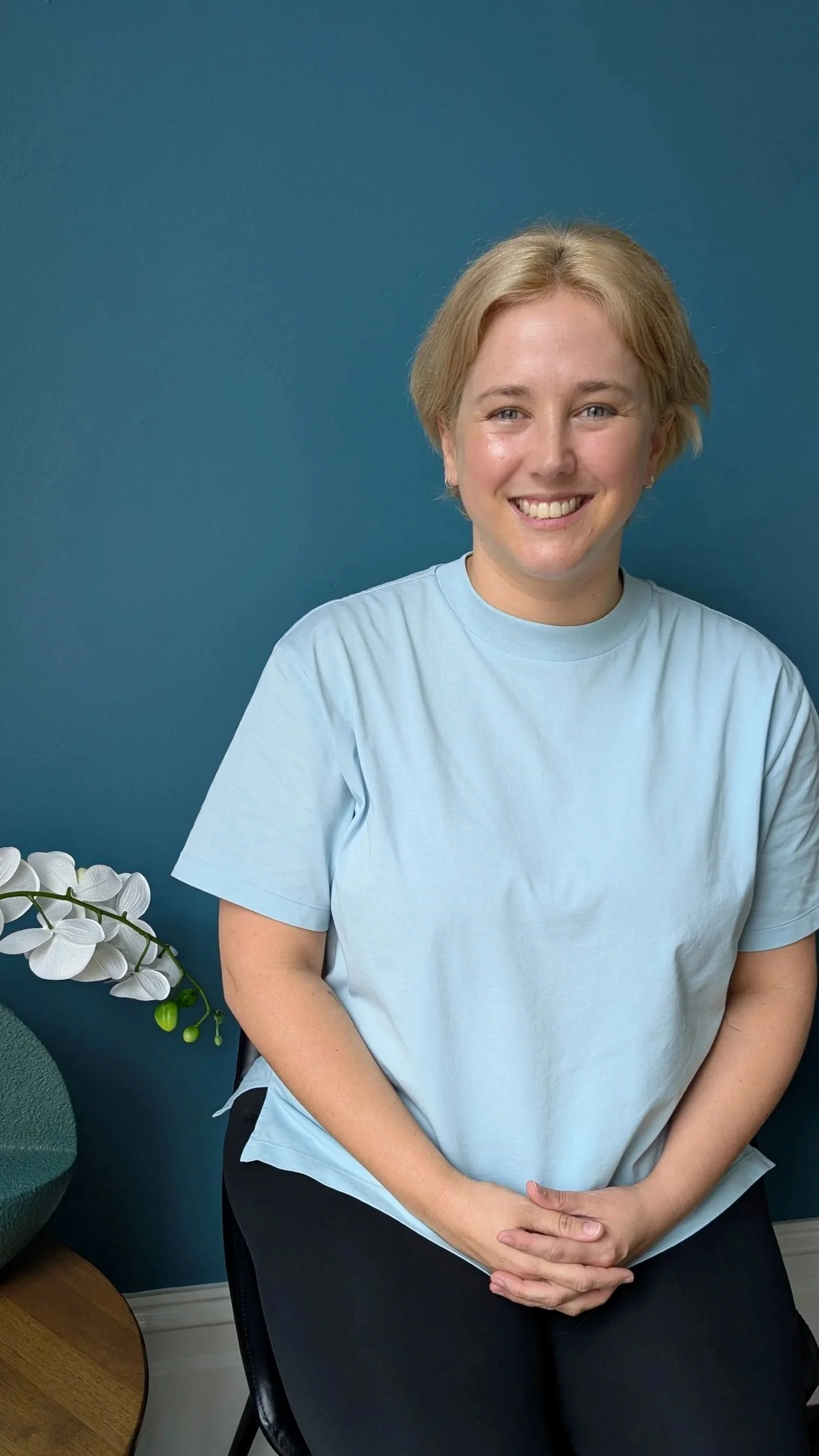 Erin is the owner of StretchWell assisted stretching, is wearing a light blue t-shirt, sitting in a black chair against a blue wall, smiling at the camera with hands folded on her lap. There is a white orchid plant on the left side.