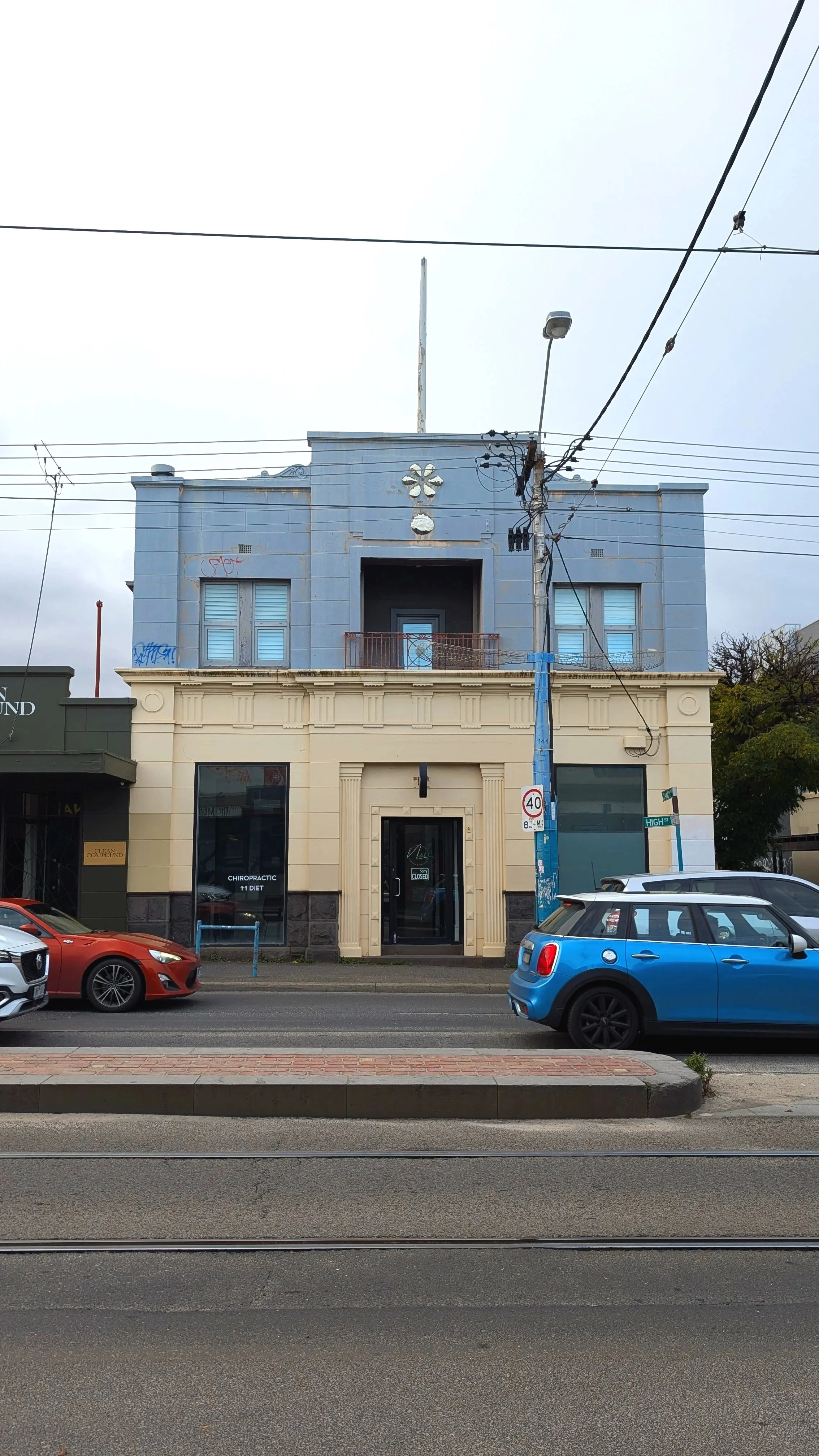 A city street scene featuring a multi-story building with a beige lower section and a blue upper section with a small balcony. Several cars are parked along the street, with tram tracks and a sidewalk in the foreground. Overhead, there are electrical wires, a streetlight, and a utility pole.