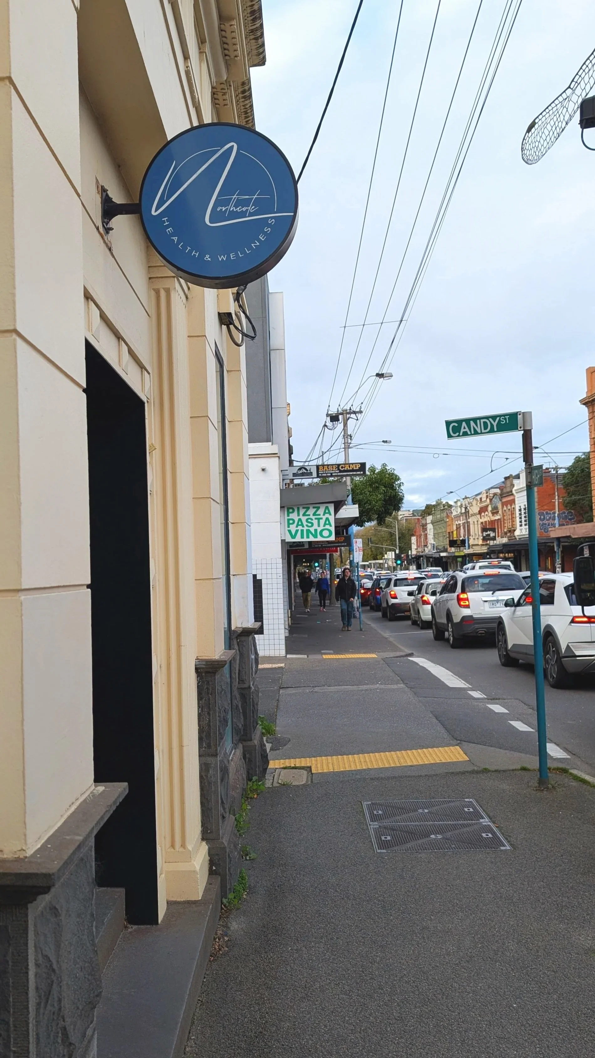 A street view with a sidewalk, storefronts, traffic, and a street sign for Candy Street. There is a sign for a health and wellness business and a sign for a restaurant serving pizza, pasta, and vino. Pedestrians are walking along the sidewalk and several cars are parked or moving on the street.