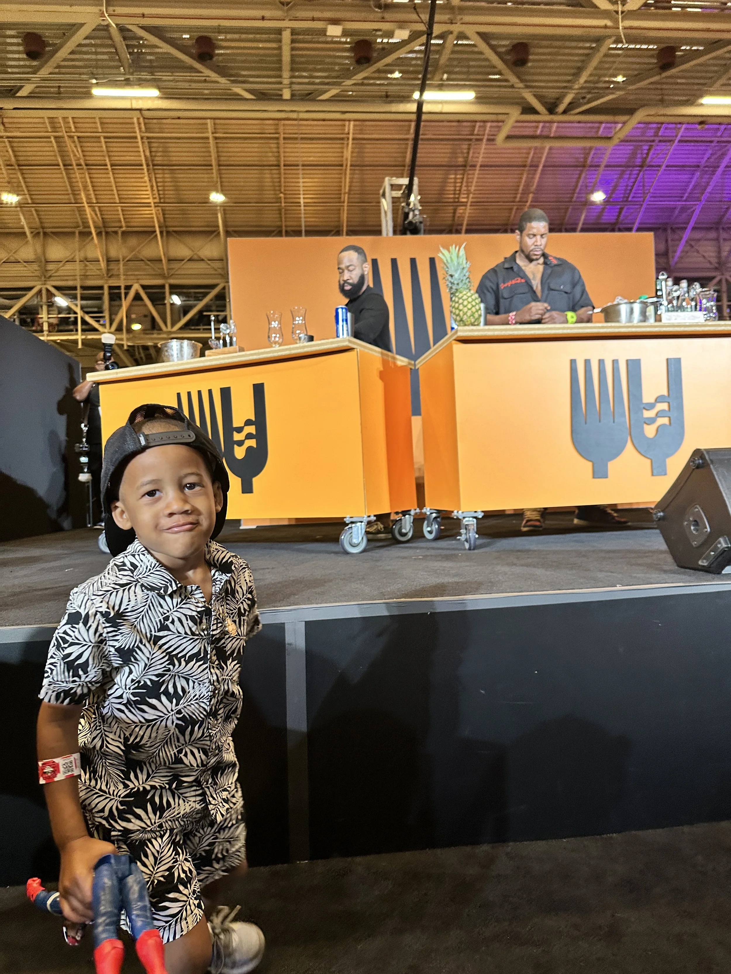 A young boy standing in front of a stage with two bartenders serving behind orange bars, one of whom has a pineapple on the counter.