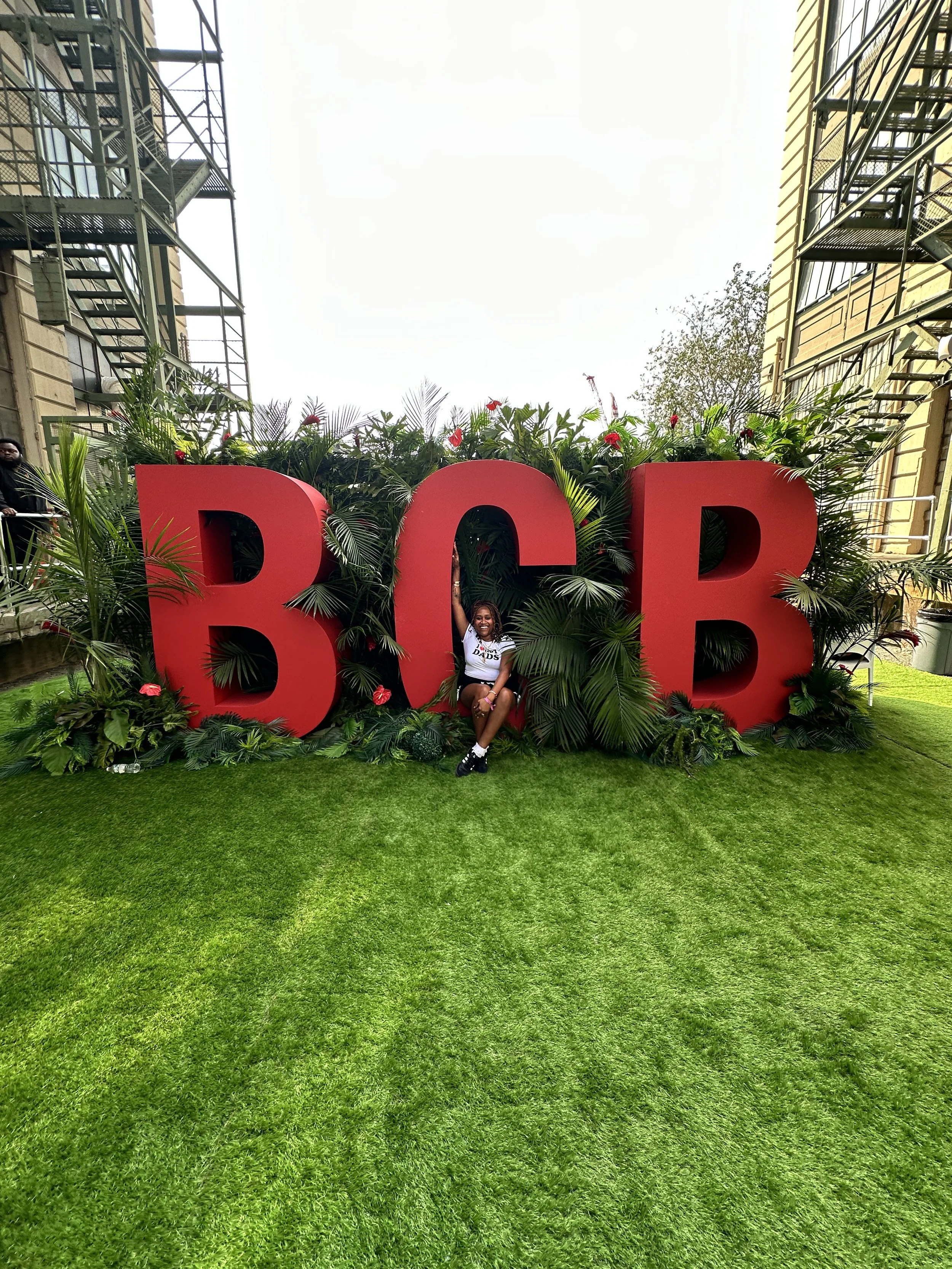 A woman sitting on the ground in front of large red letters spelling 'BCB', with green tropical plants around the letters and a grassy area in the foreground, in an urban setting with buildings and scaffolding nearby.