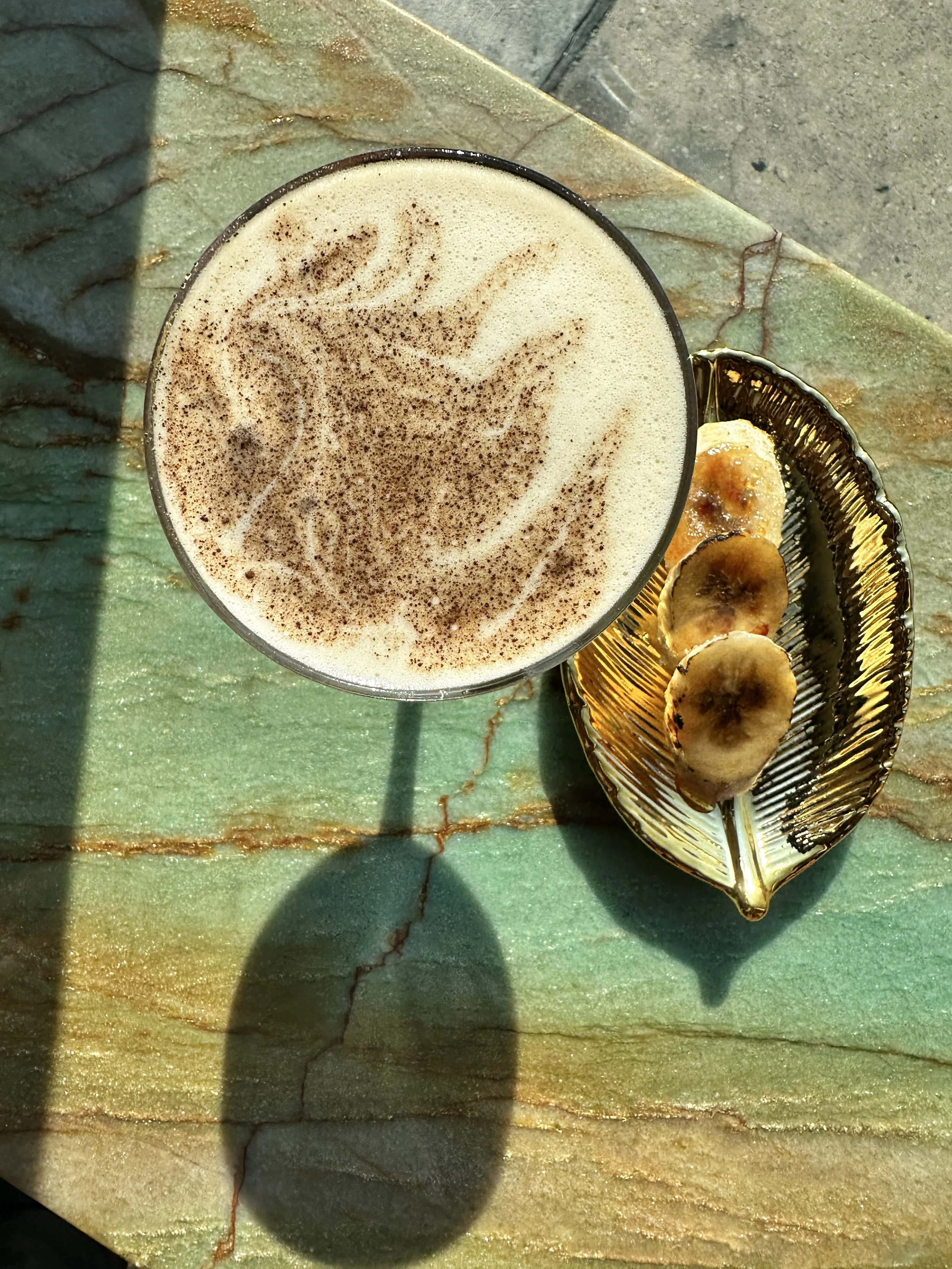A glass of frothy coffee topped with cinnamon, next to a golden dish with sliced bananas on a marble surface.