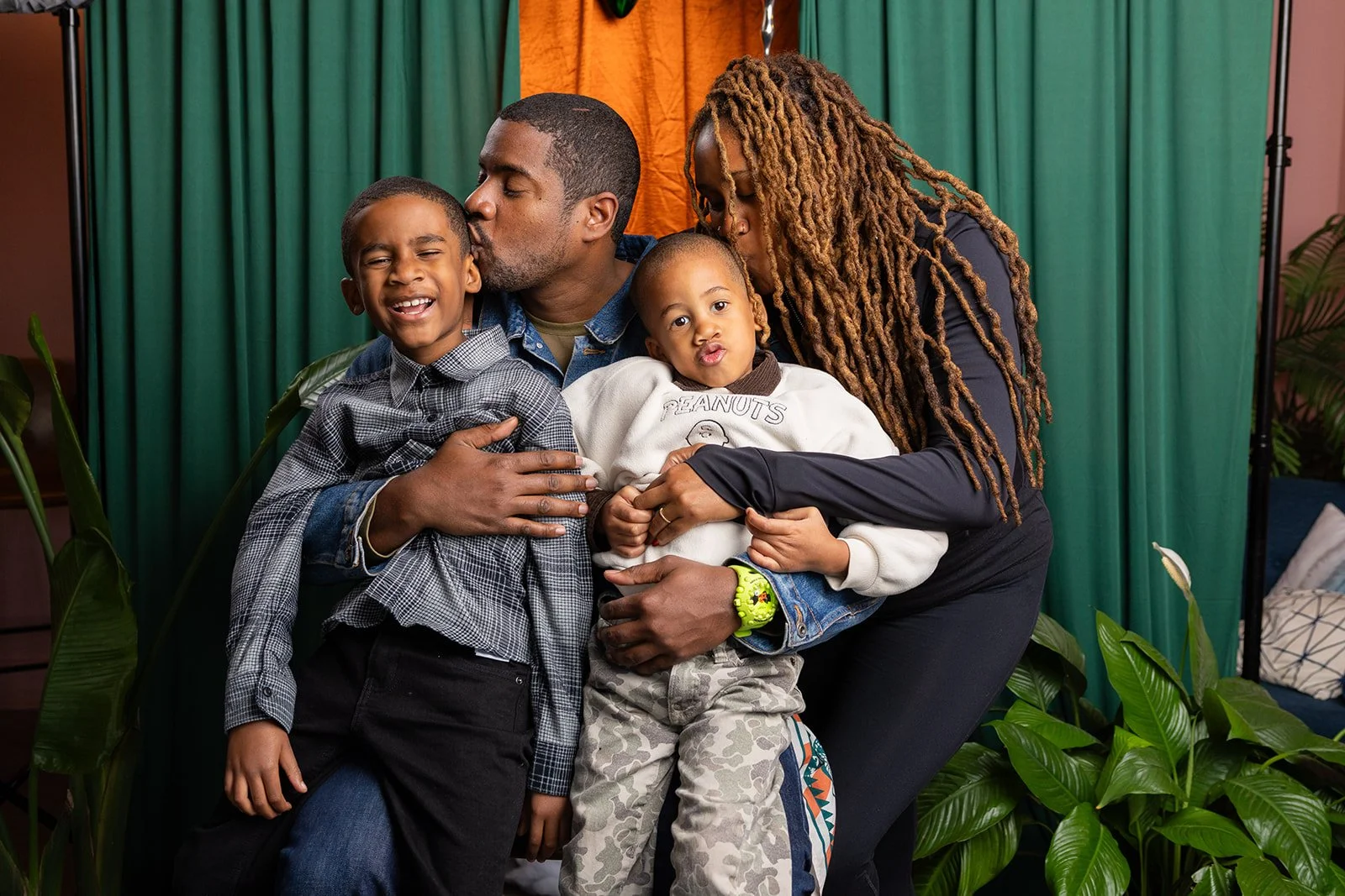 A family of four, including a man, woman, and two young boys, sharing an affectionate moment on a stage with a green curtain backdrop and plants in the foreground.