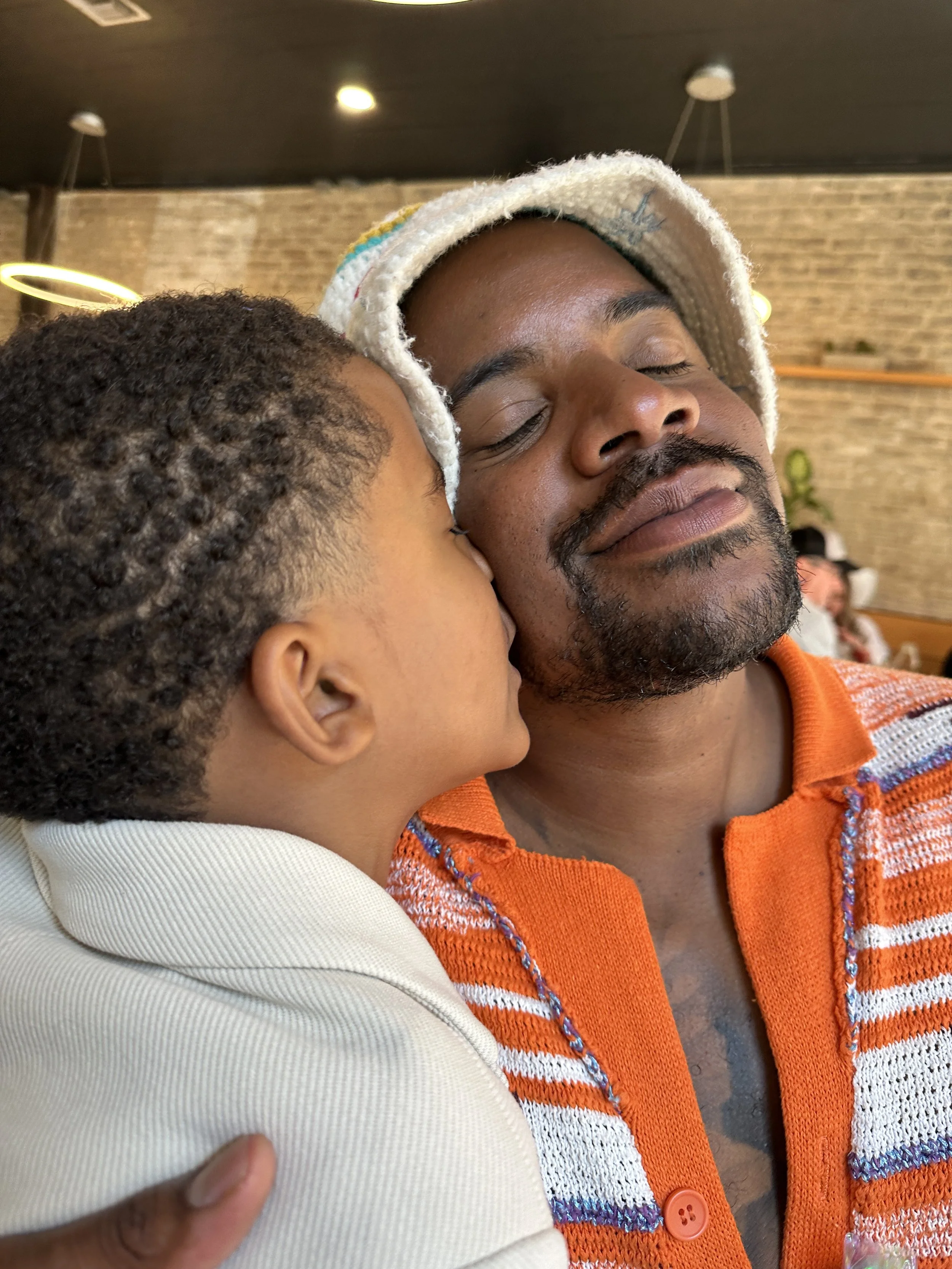 A child hugs a man, kissing his cheek, both with closed eyes, inside a cafe with a brick wall background.