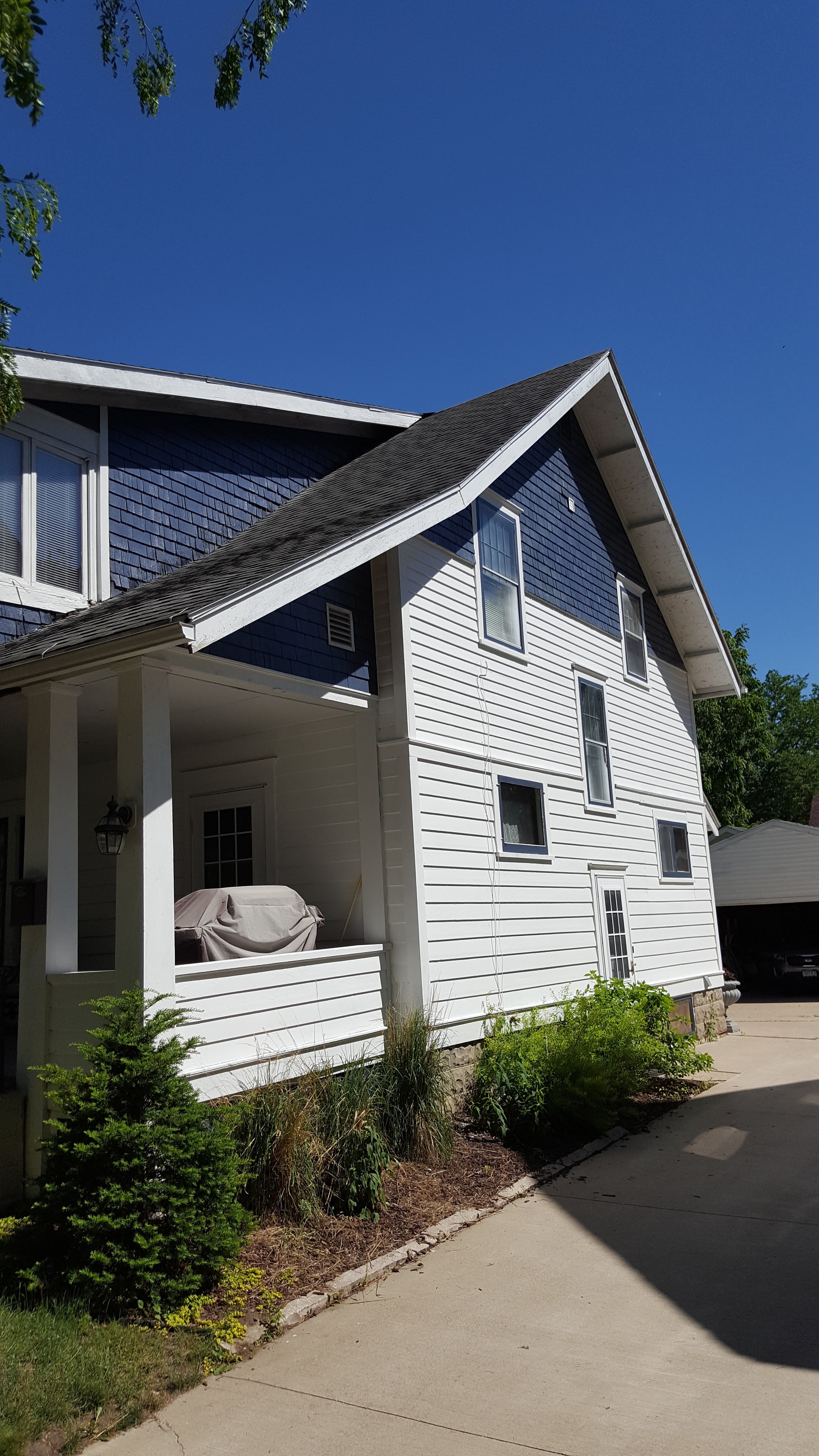 Two-story house with white siding, multiple windows, a porch with a covered grill, and a driveway with a parked car nearby. The house has a dark blue section with shingles and a sloped roof, under a clear blue sky.