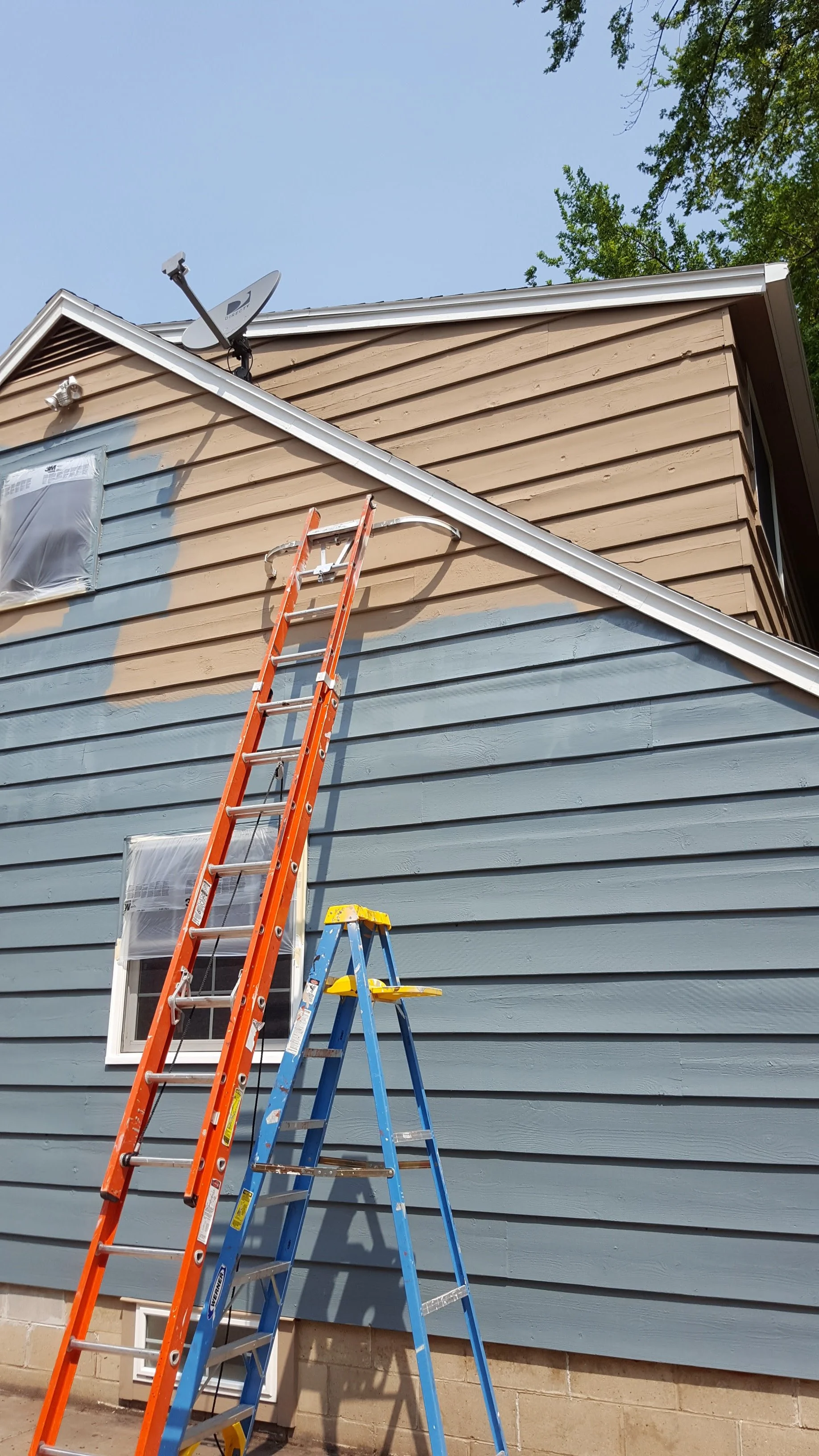 Ladders leaning against the side of a house with blue and tan siding. A satellite dish and two small covered windows are visible on the house. The sky is clear and there are green trees overhead.
