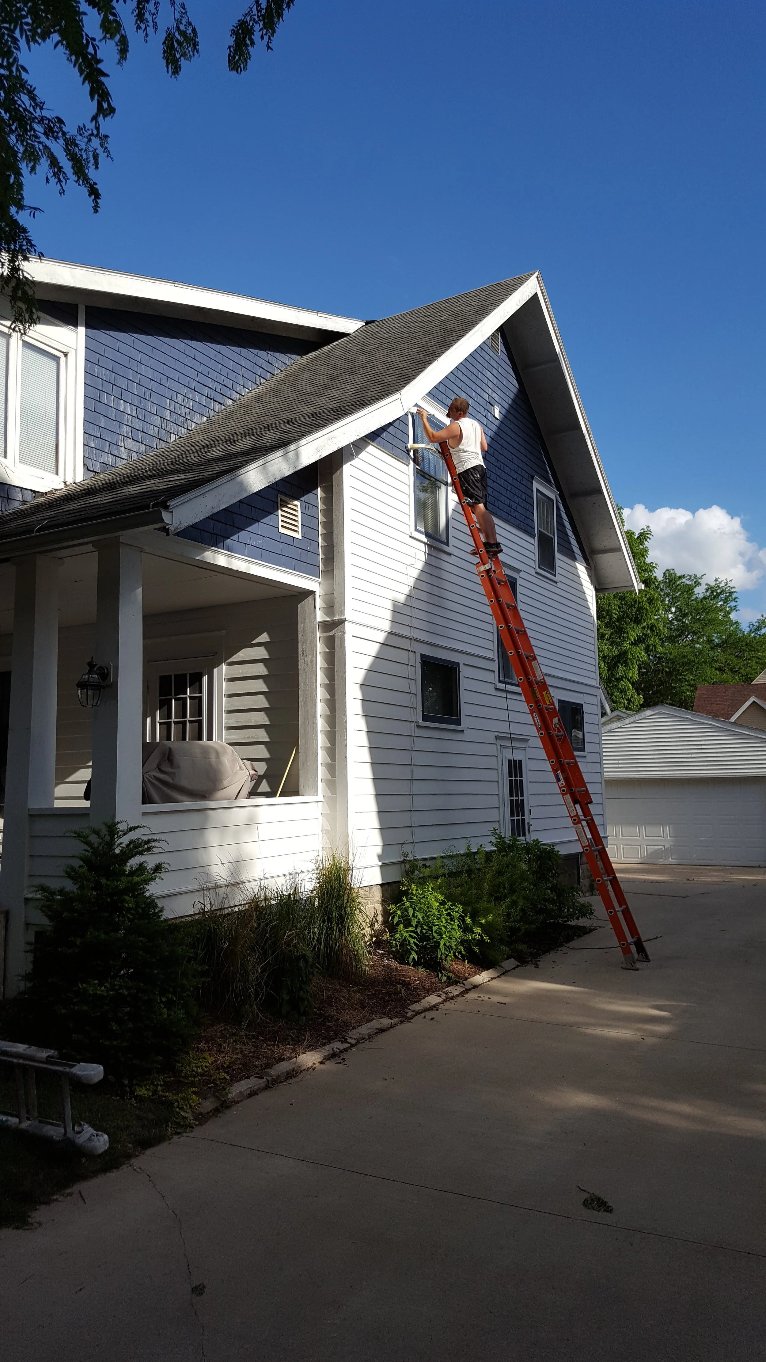 A person standing on an extension ladder cleaning or inspecting the exterior window of a two-story house with blue and white siding. The house has a front porch and a driveway, with a garage in the background. The sky is clear and blue.