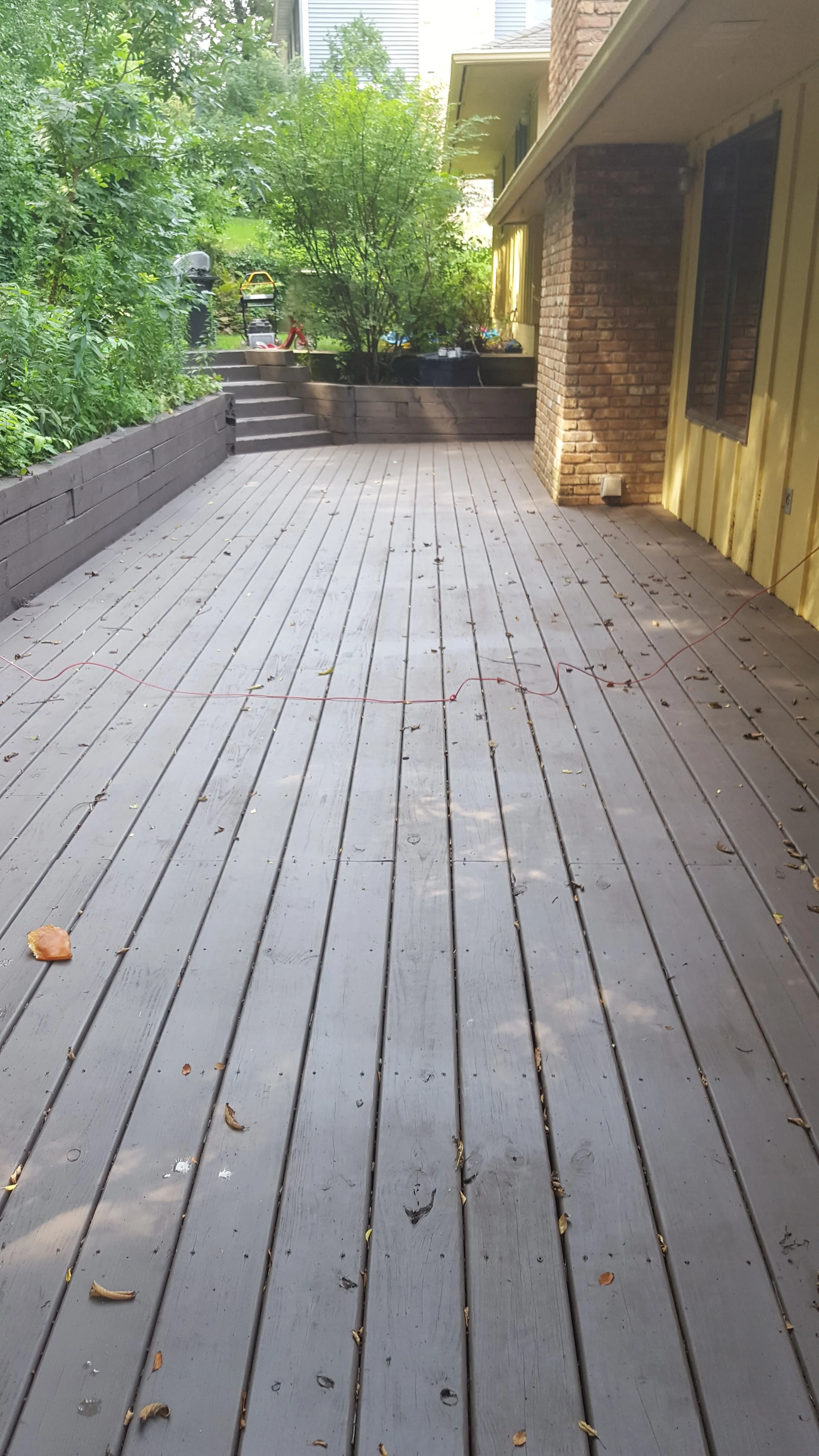 A wooden deck outdoor patio with scattered leaves and twigs, adjacent to a brick and yellow siding house, with lush green trees and plants in the background. A red string or cord is lying across the deck.