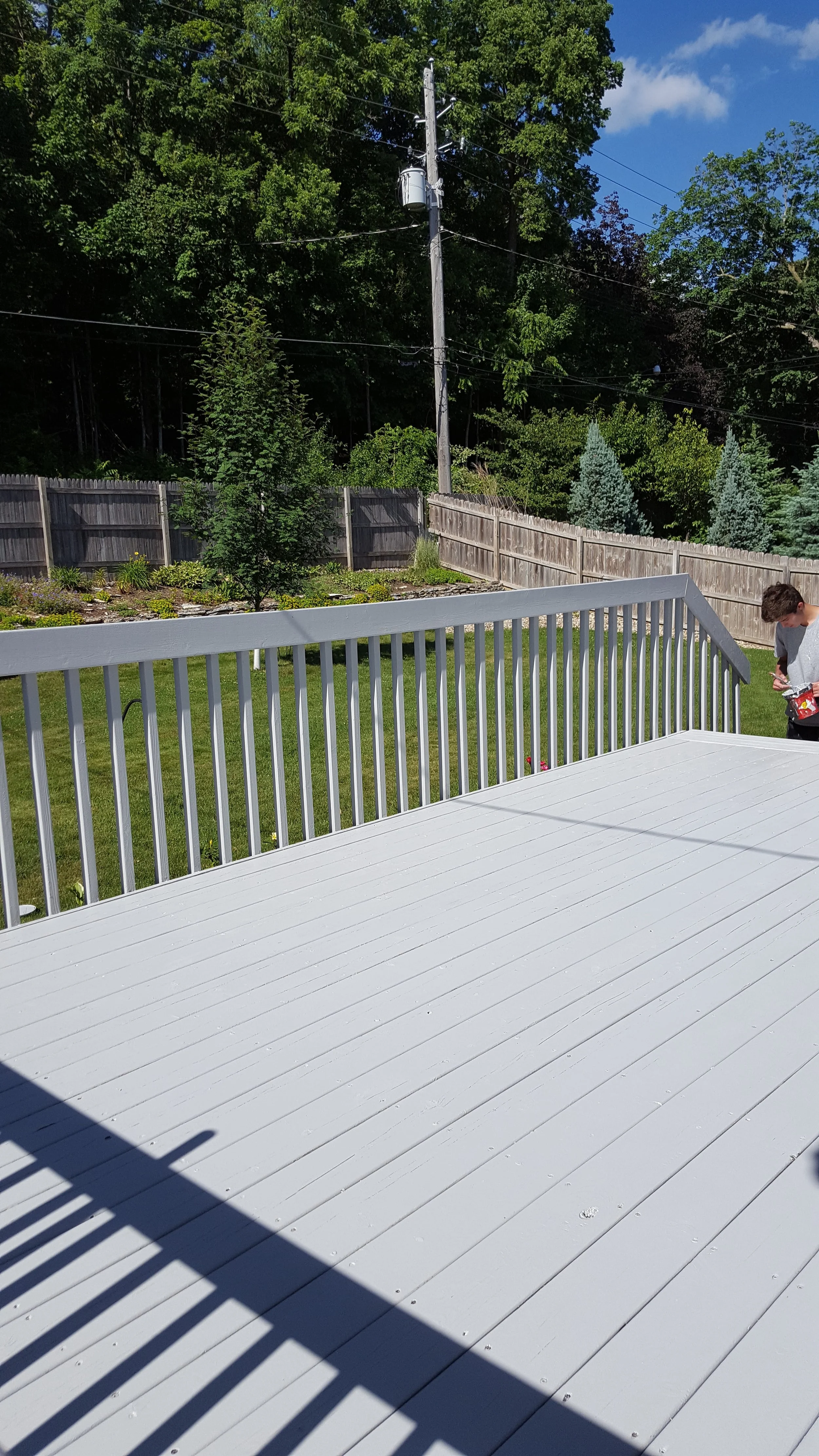 Clean white wooden deck with a railing, outdoor backyard garden with green grass, trees, and a wooden fence under a partly cloudy sky. A person in a gray shirt is visible in the corner, holding a red object.