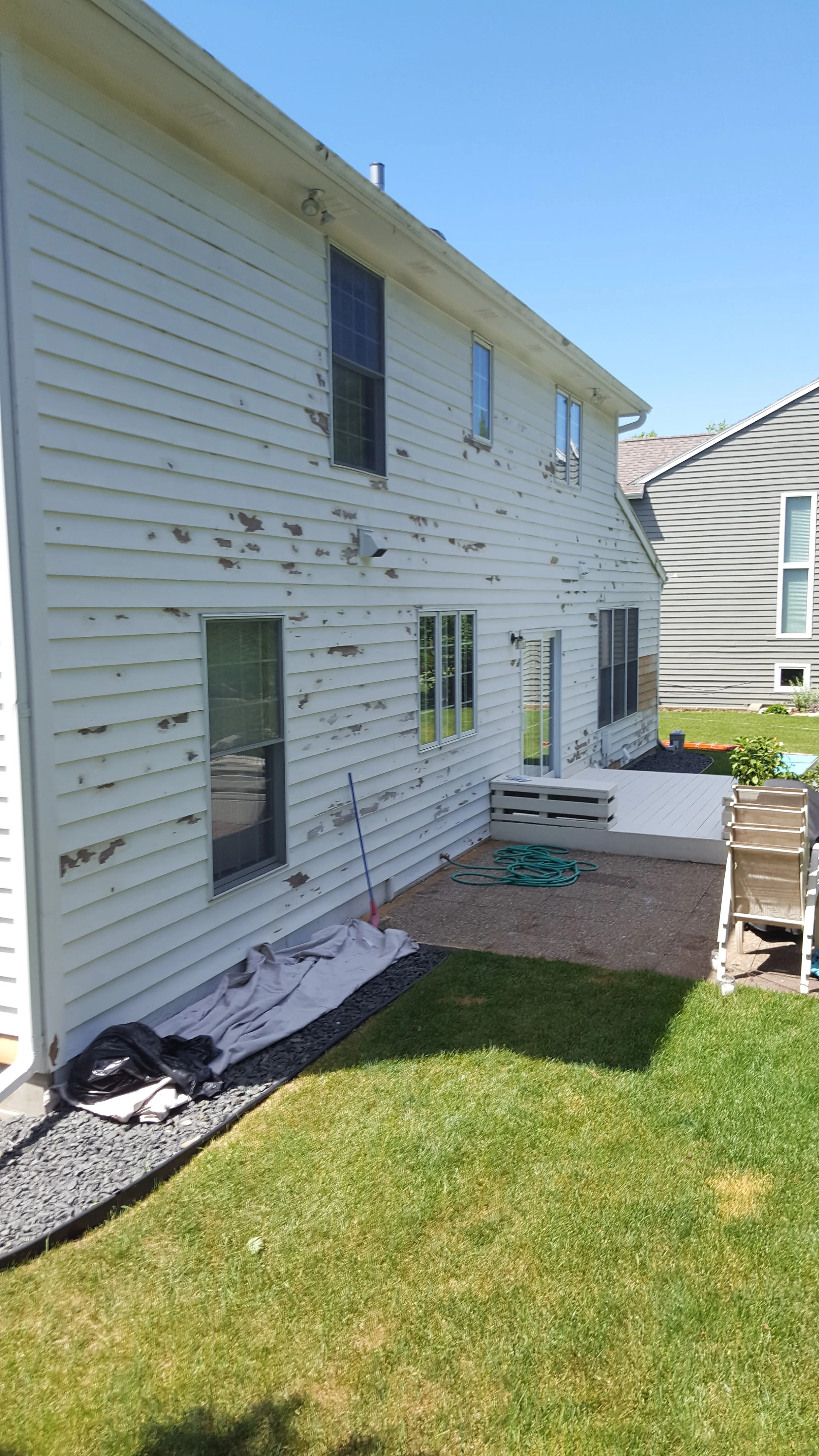 Backyard of a house with white siding showing peeling paint, a small deck, green lawn, and a neighboring house in the background.