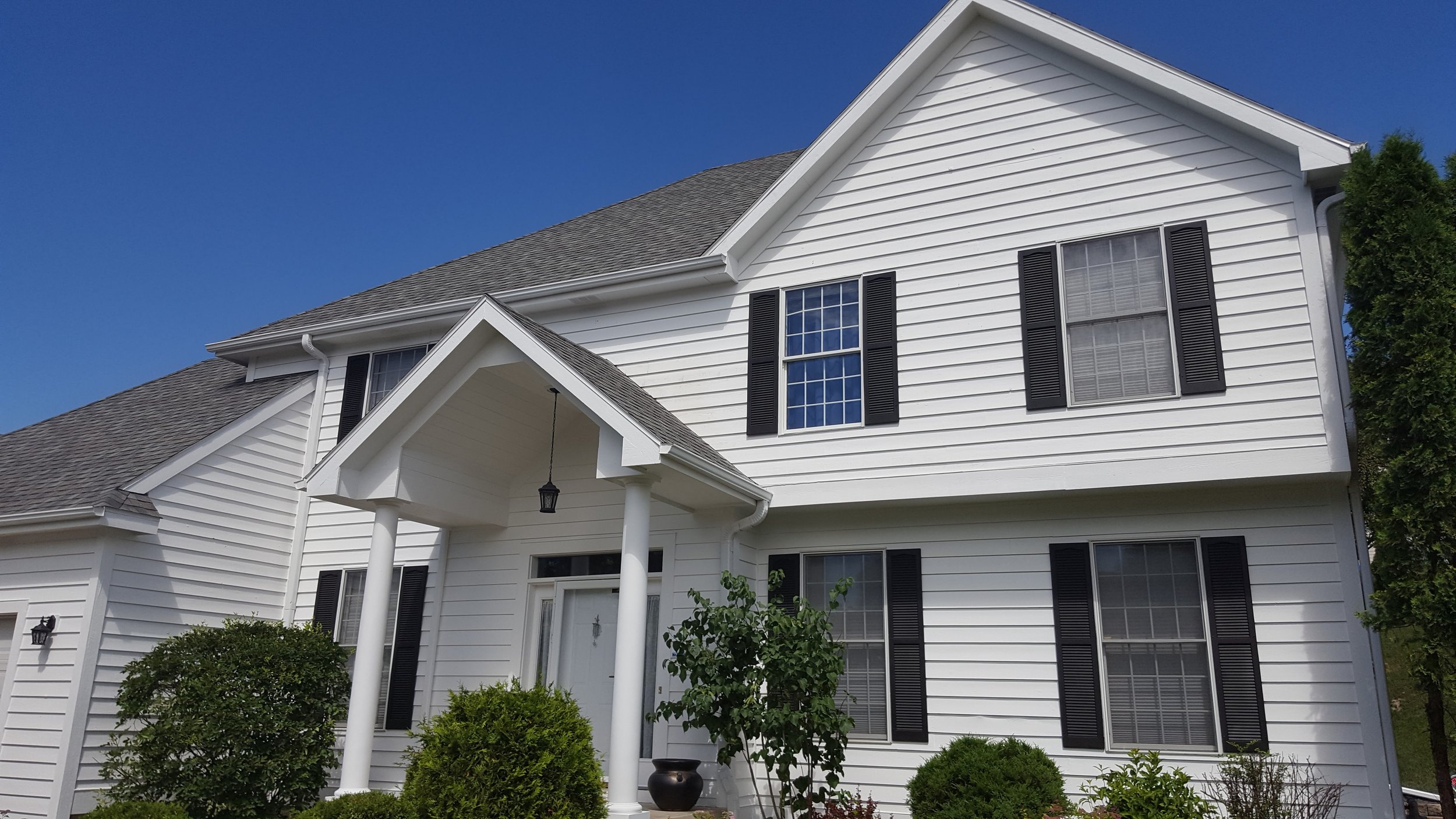 White two-story house with black shutters, front porch with columns, potted plant, bushes, and a clear blue sky.