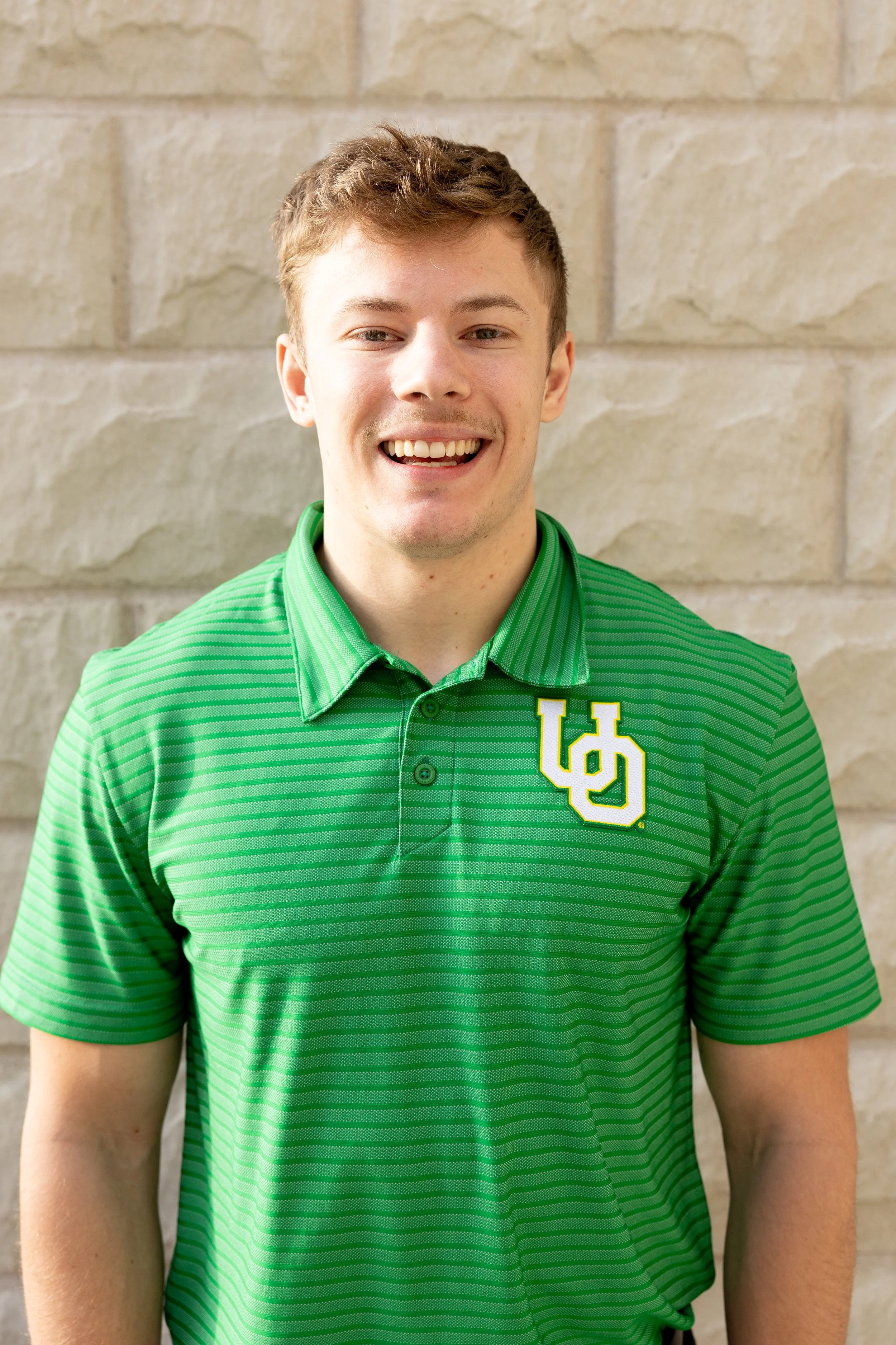 A young man with short brown hair smiling, wearing a green striped polo shirt with a yellow and white logo on the chest, standing in front of a beige brick wall.