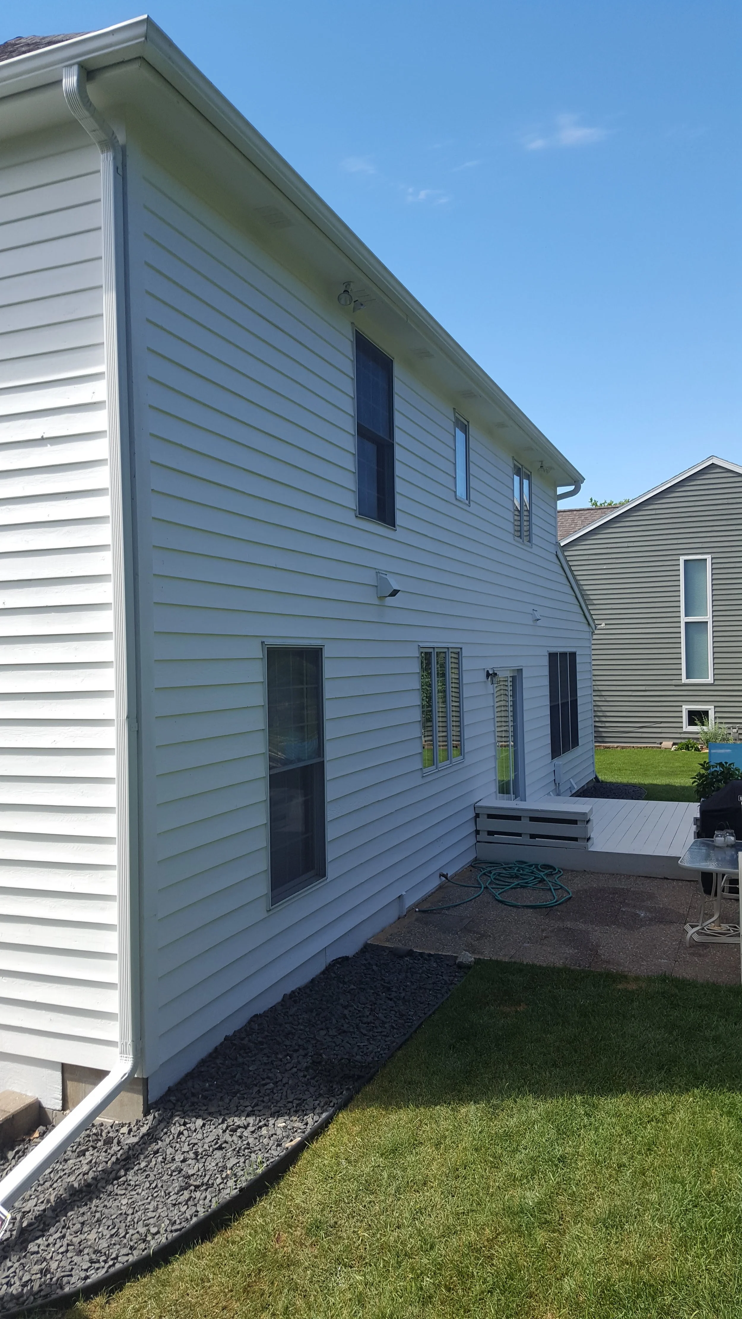 Side view of a white, two-story house with multiple windows, a small porch, a garden hose, and a patio area, with green grass and a neighboring house visible in the background under a blue sky.