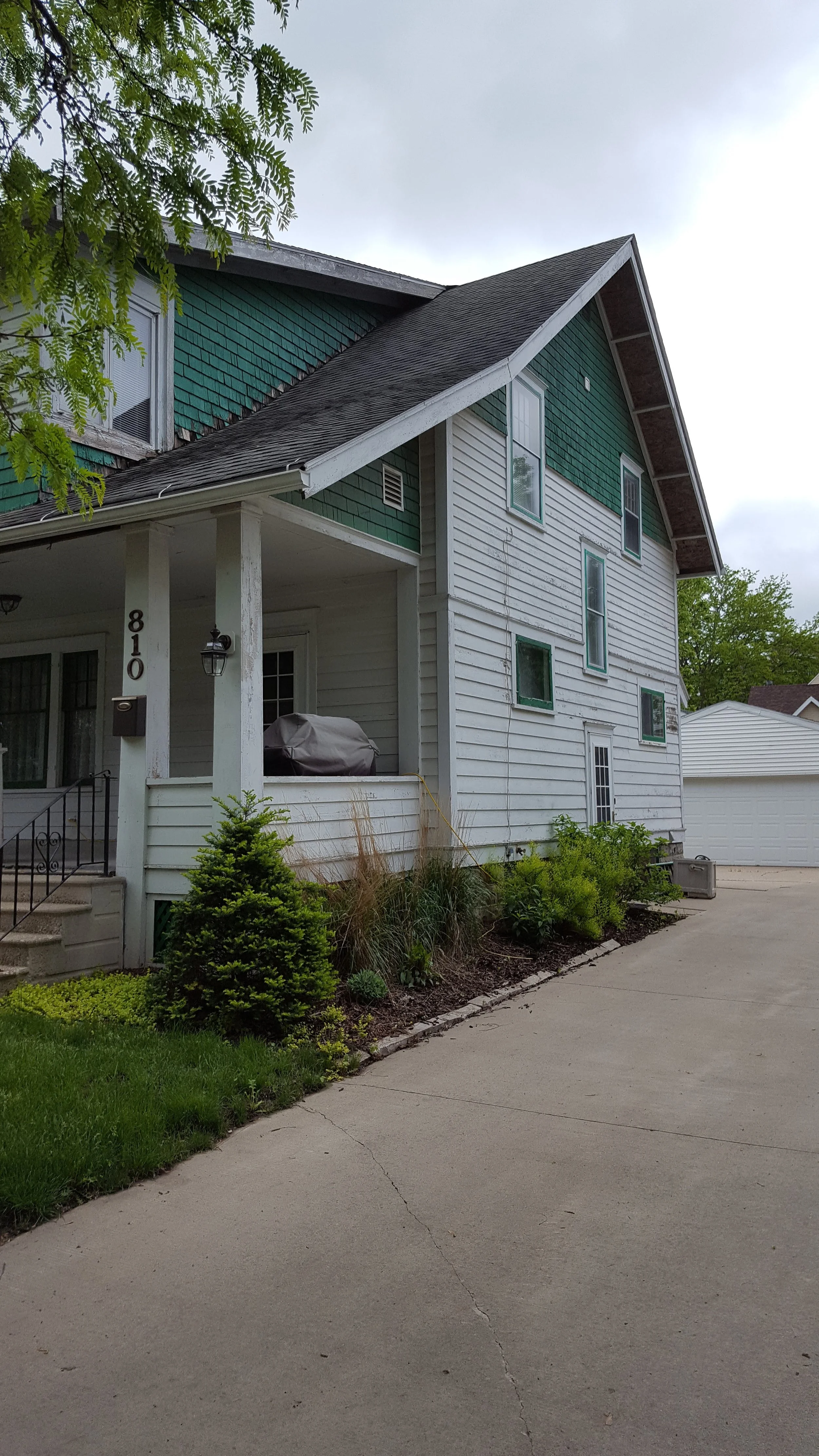 A two-story house with green and white siding, small front porch, stairs, and a driveway with some plants and shrubs along the side.