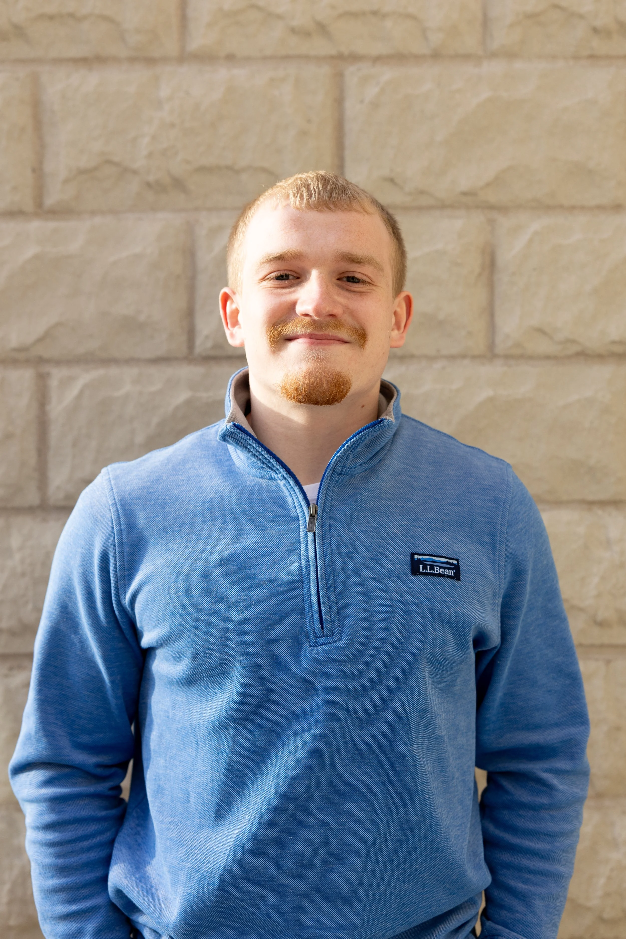 Portrait of a young man with light skin, short light brown hair, and a mustache and goatee, wearing a blue quarter-zip fleece jacket standing against a beige brick wall.