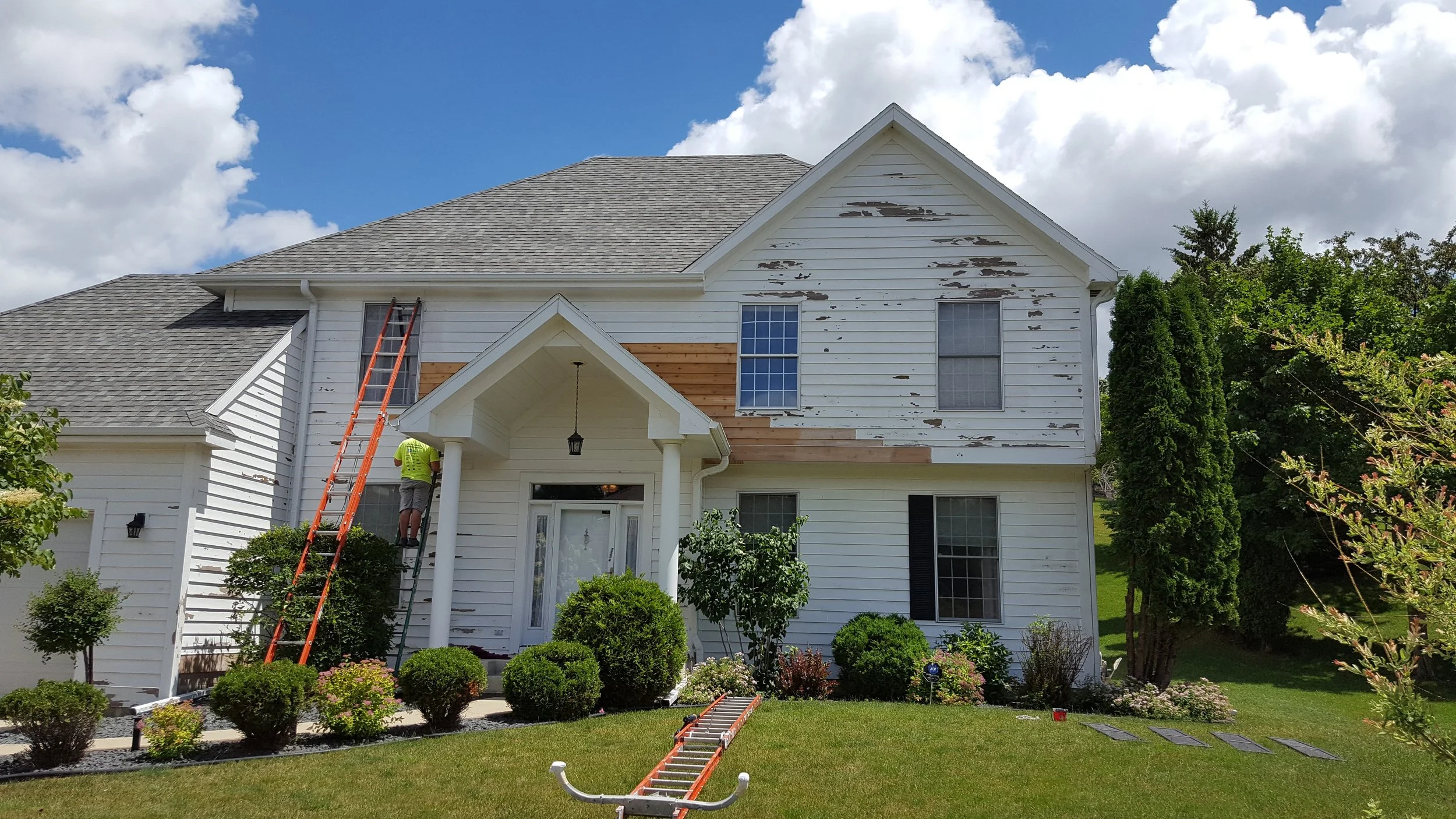 Two-story white house with damage to the siding, workers on ladders repairing it, and landscaping with bushes and trees in front.