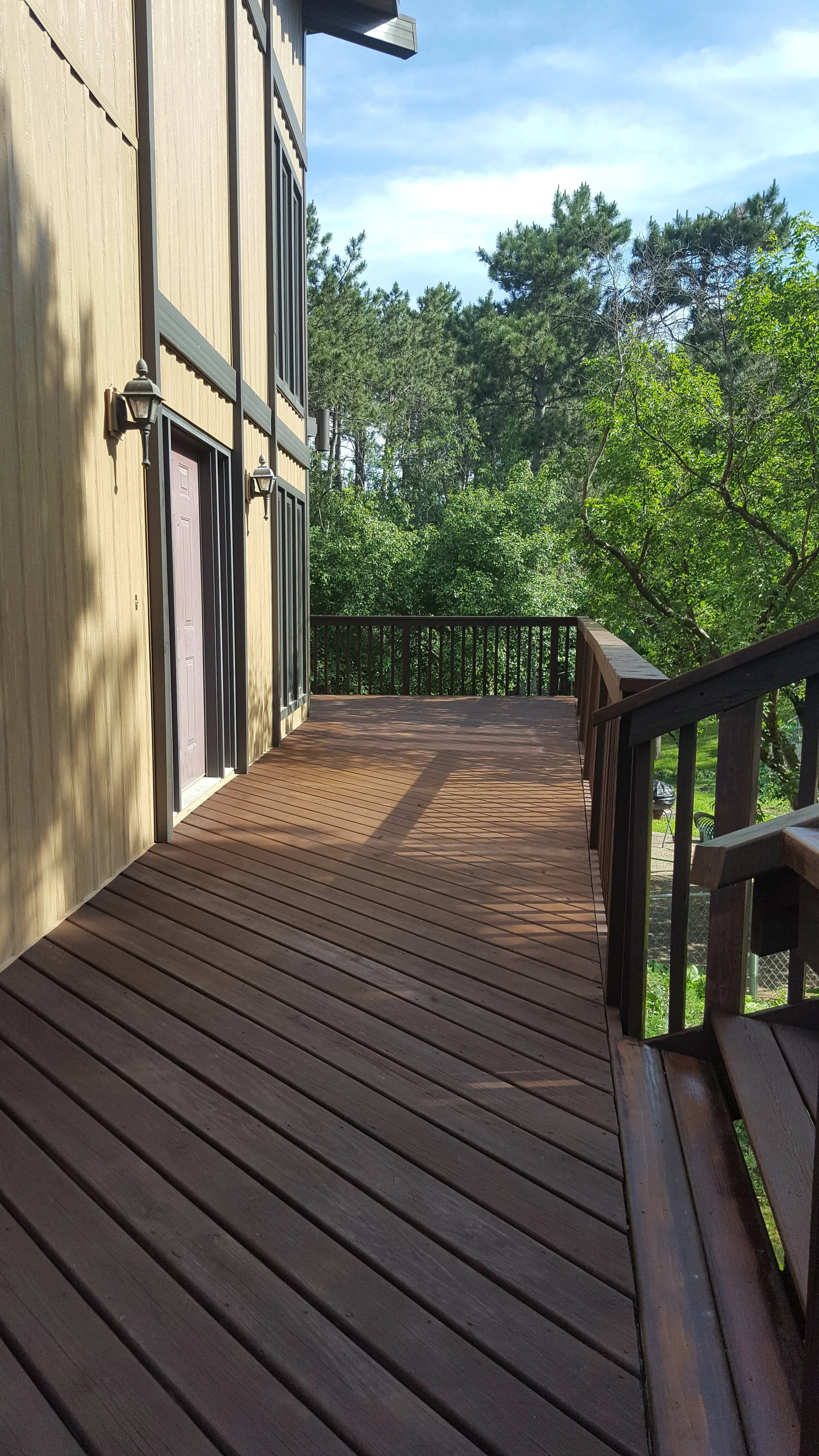 Wooden deck attached to a house with two doors, outdoor lights, surrounded by green trees and blue sky.