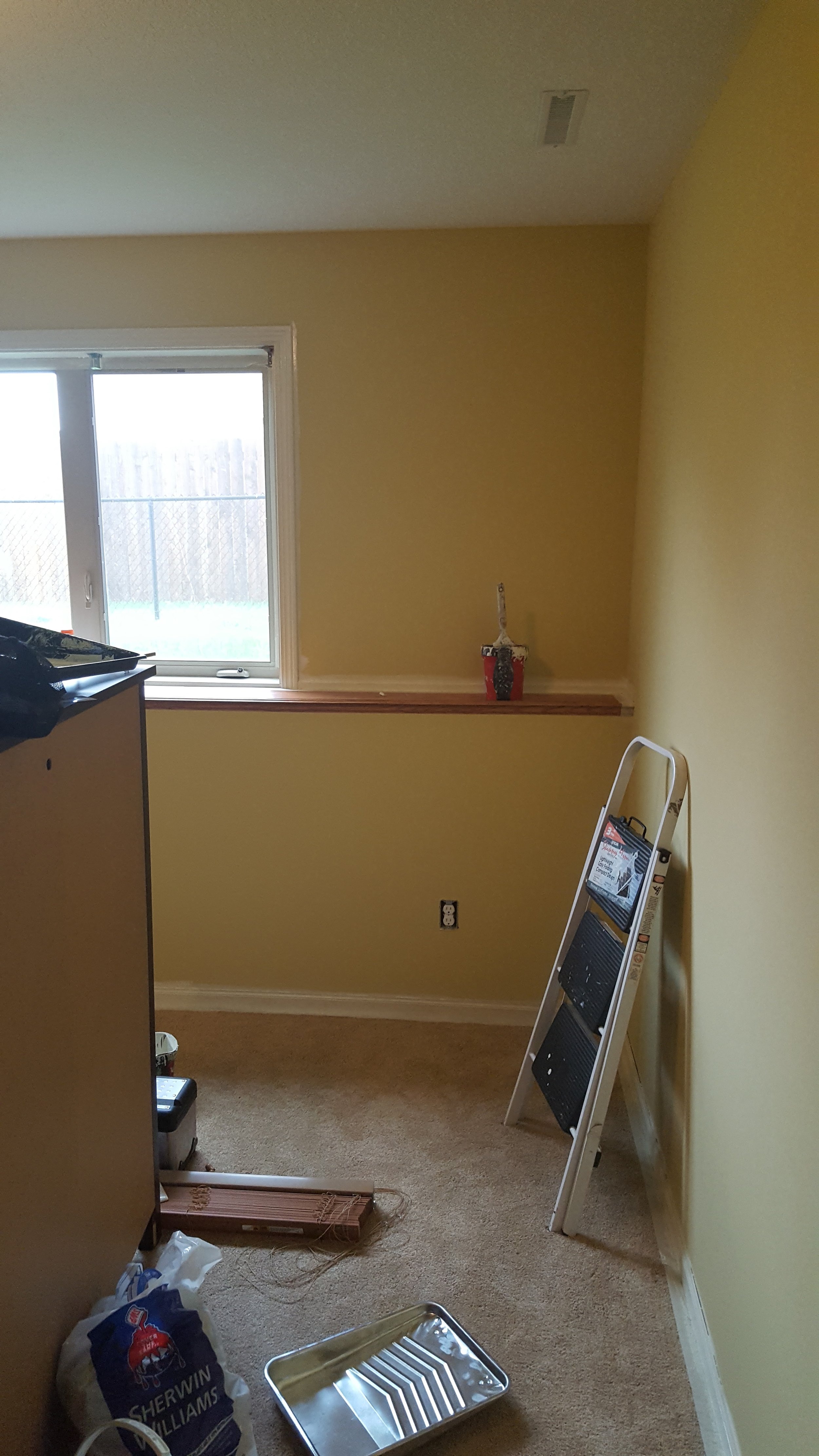 A small, empty room with beige carpet, yellow walls, and a large window. There is a step ladder leaning against the wall, a small tray on the floor, and a bag of Sherwin-Williams paint near the bottom left corner. A narrow wooden ledge runs along the