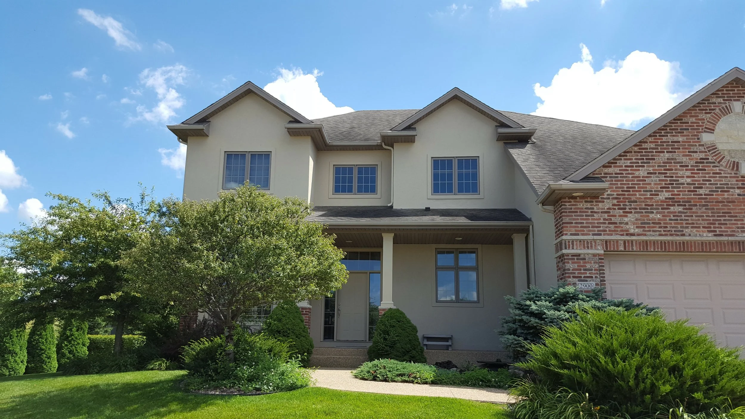 A two-story house with a beige and brick exterior, a front porch, and a well-manicured lawn with bushes and trees under a partly cloudy sky.