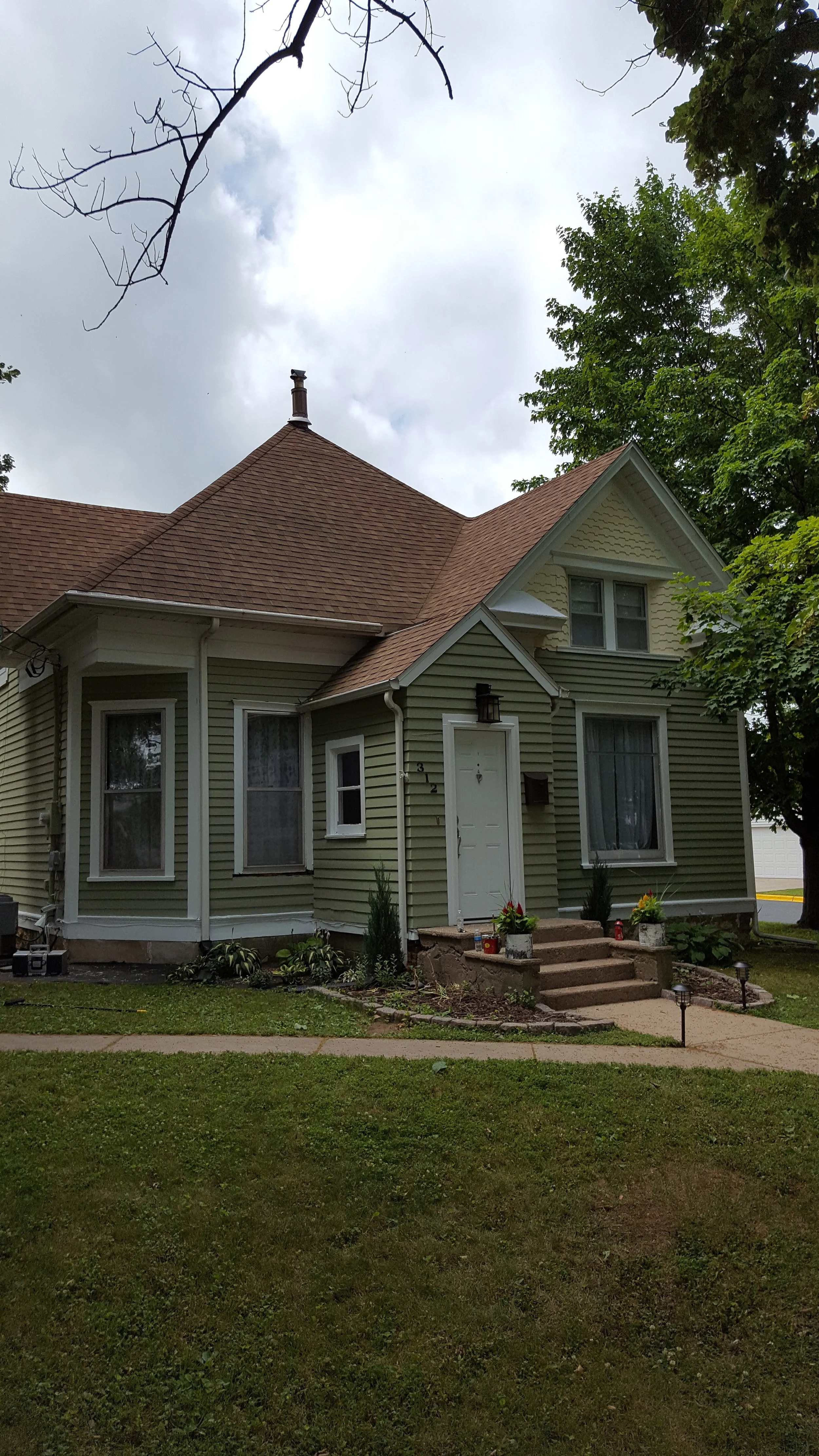 A two-story house with light green siding, a brown shingled roof, and a small front porch with steps. There are plants and flowers near the entrance, and a sidewalk leading to the house, with trees around.