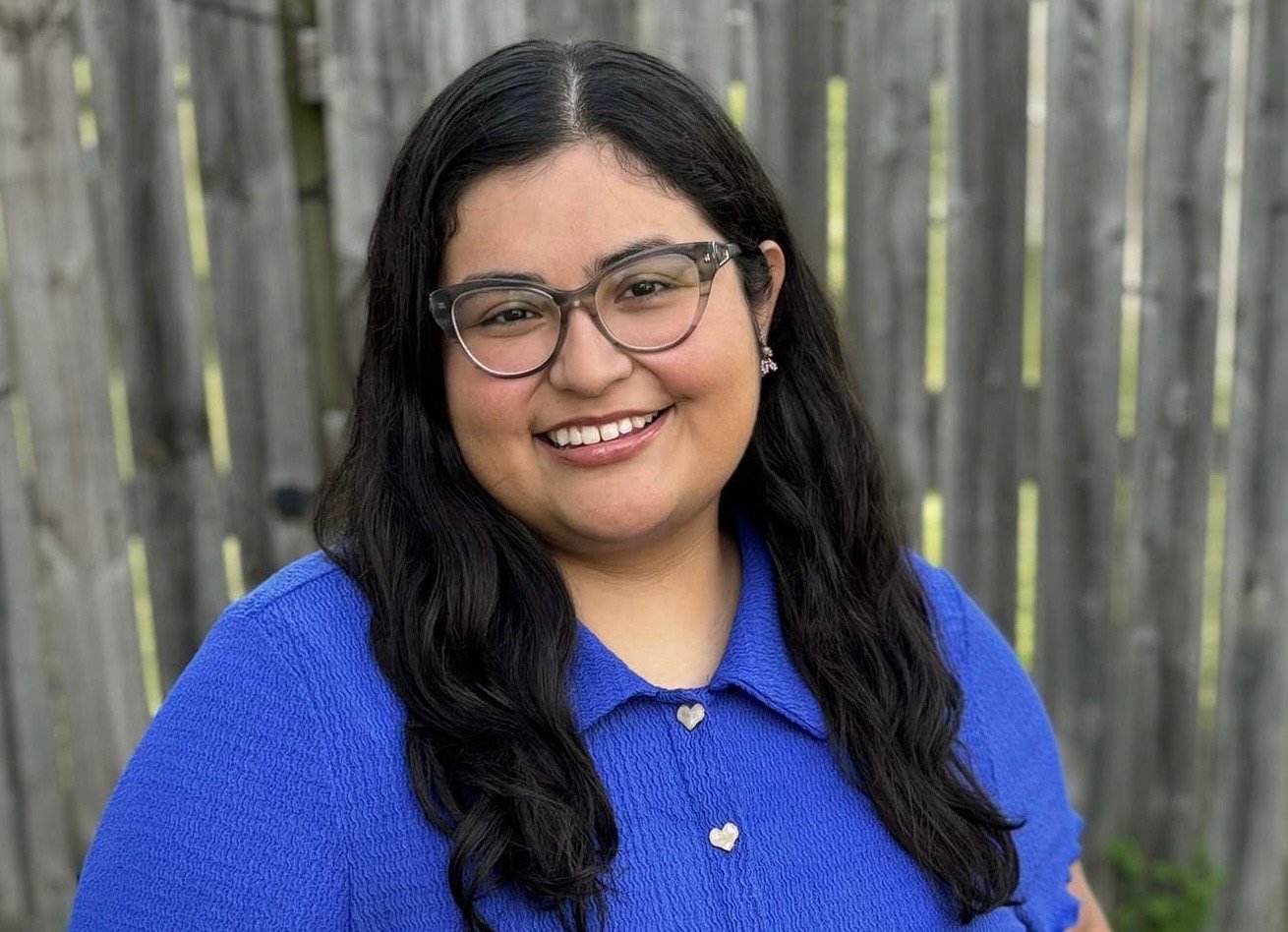 A woman with long dark hair, wearing glasses and a blue shirt, smiling outdoors in front of a wooden fence.