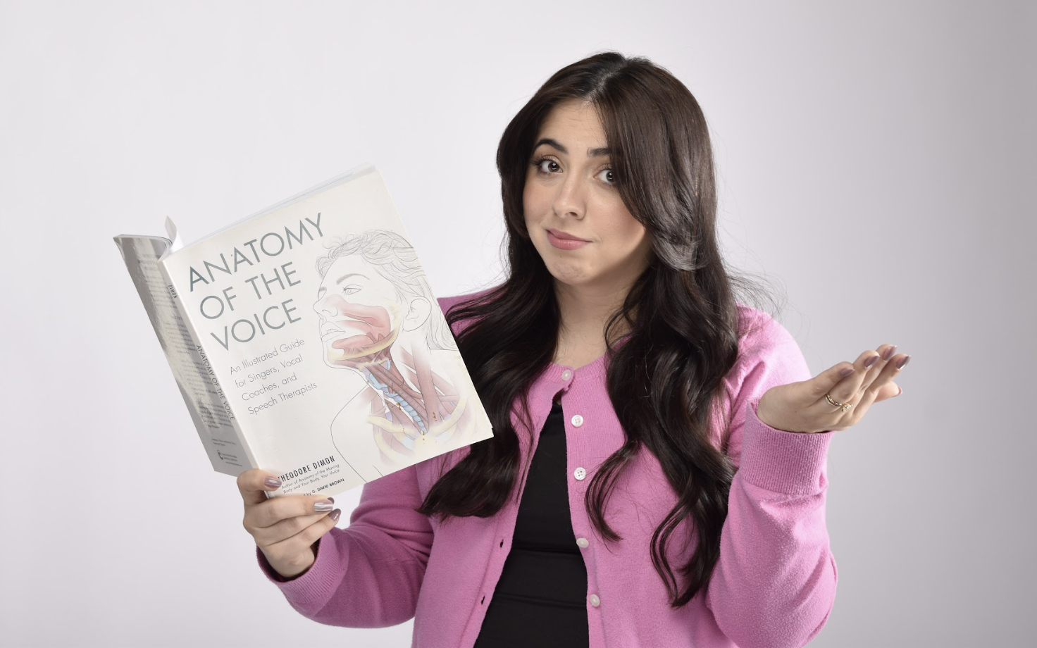 A woman holding a book titled 'Anatomy of the Voice' with a diagram of the vocal anatomy, wearing a pink cardigan and black shirt, standing against a plain background.