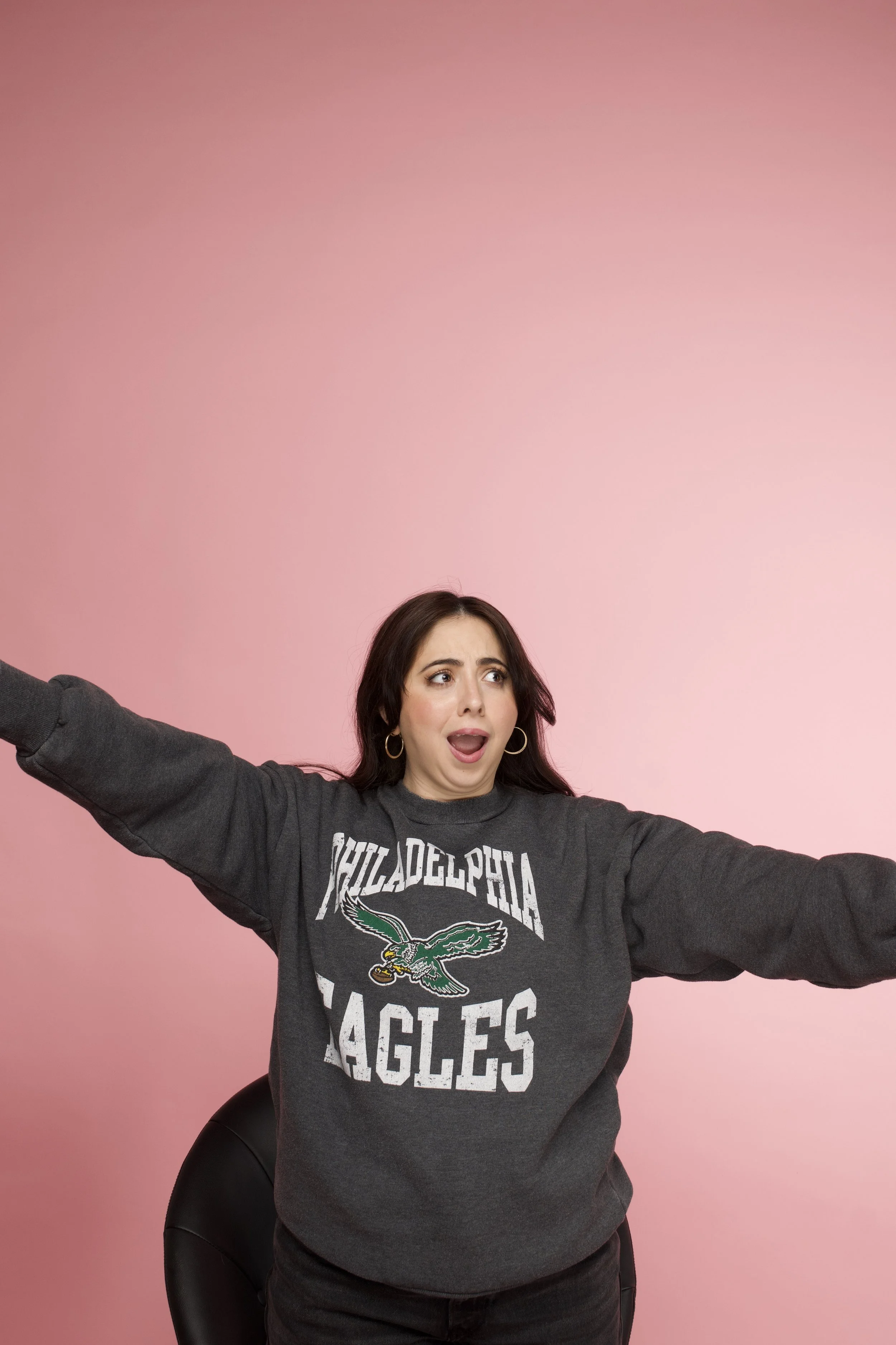 A woman with dark hair wearing a dark grey Philadelphia Eagles sweatshirt and black pants, sitting on a black chair with a pink background, making a surprised or excited facial expression with arms stretched out.