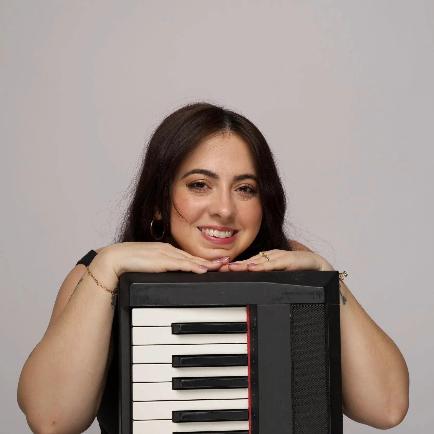 A woman with dark brown hair and hoop earrings, resting her chin on her crossed arms, holding a small keyboard in front of her face, smiling at the camera against a plain light grey background.