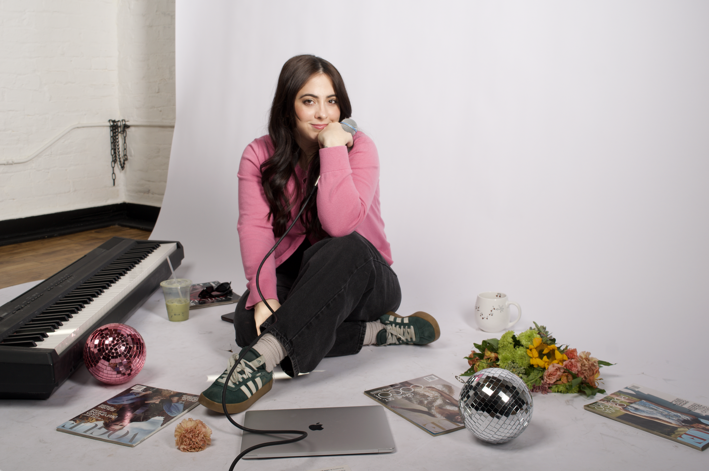 A young woman with long brown hair, wearing a pink sweater, sits on the floor with her knees up, resting her chin on her hand while holding a microphone. She is surrounded by a keyboard, magazines, flower bouquets, disco balls, a computer, a drink, and other items in a photography studio with a white backdrop.