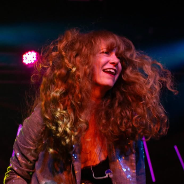 A woman with long, curly red hair smiling on stage under pink and purple lighting.