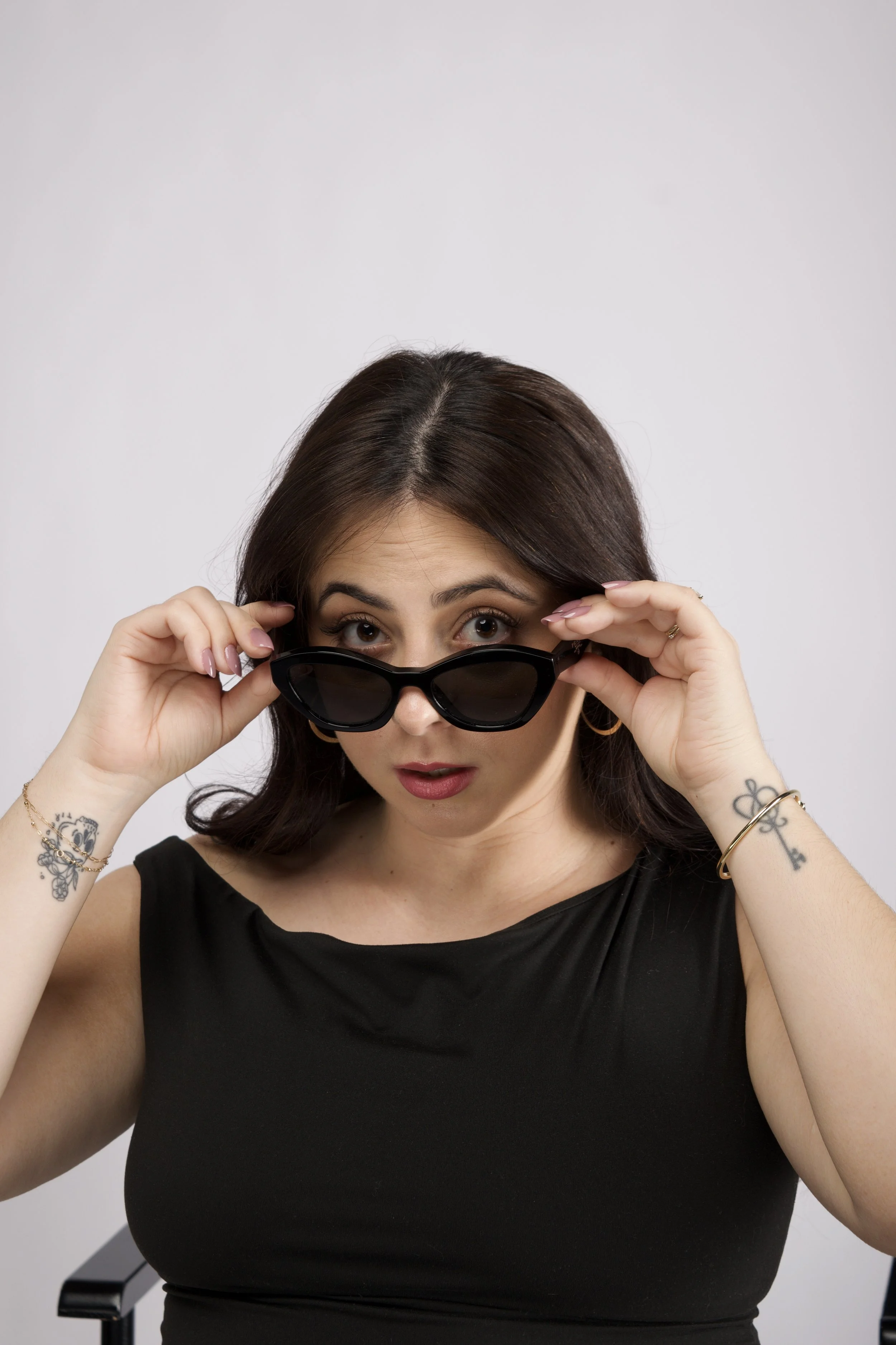 Woman with dark brown hair, holding sunglasses and looking at the camera, wearing a black sleeveless top, with tattoos on her left arm and wrist, against a plain light gray background.