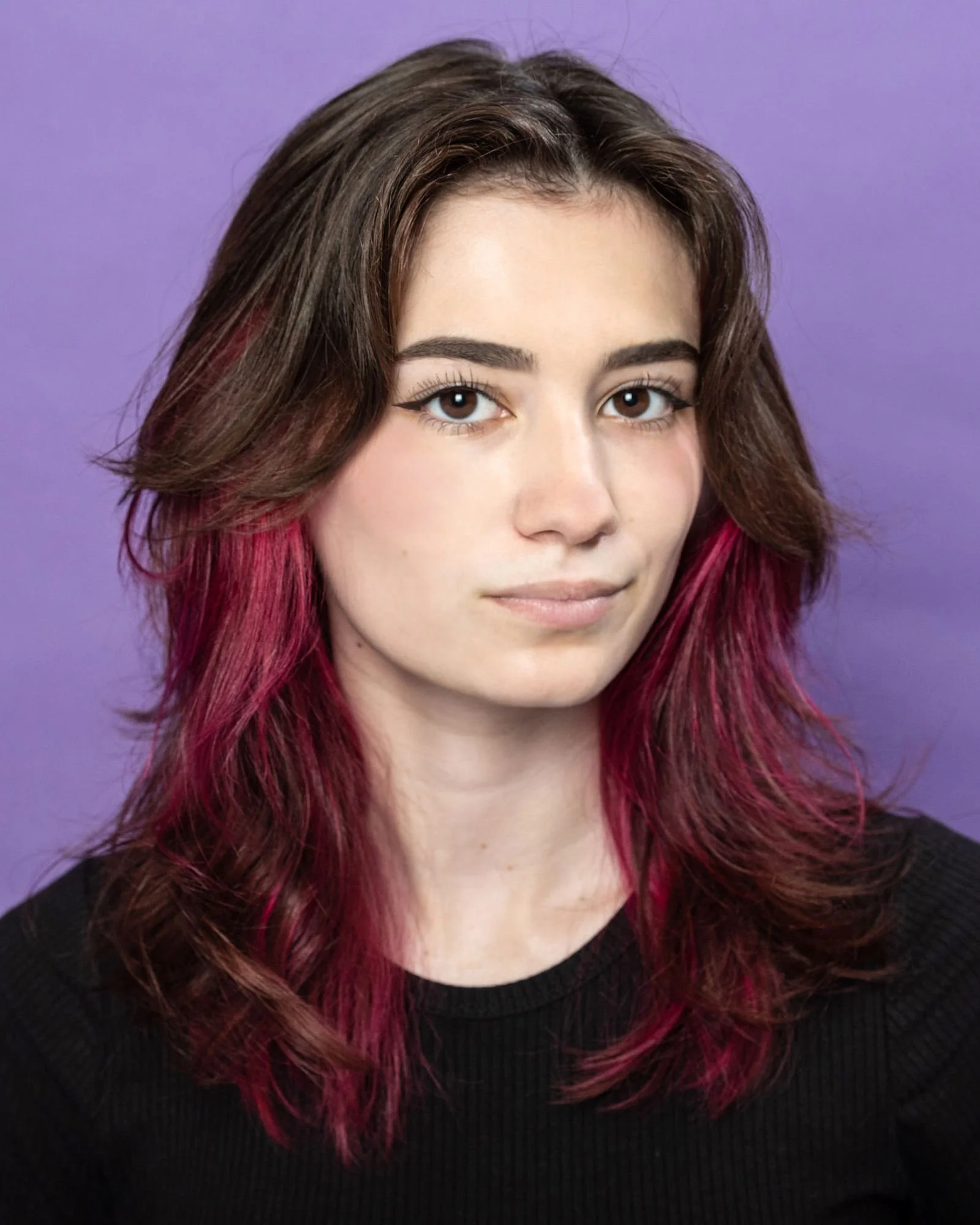 Close-up of a woman with brown and pink hair wearing a black top against a purple background.