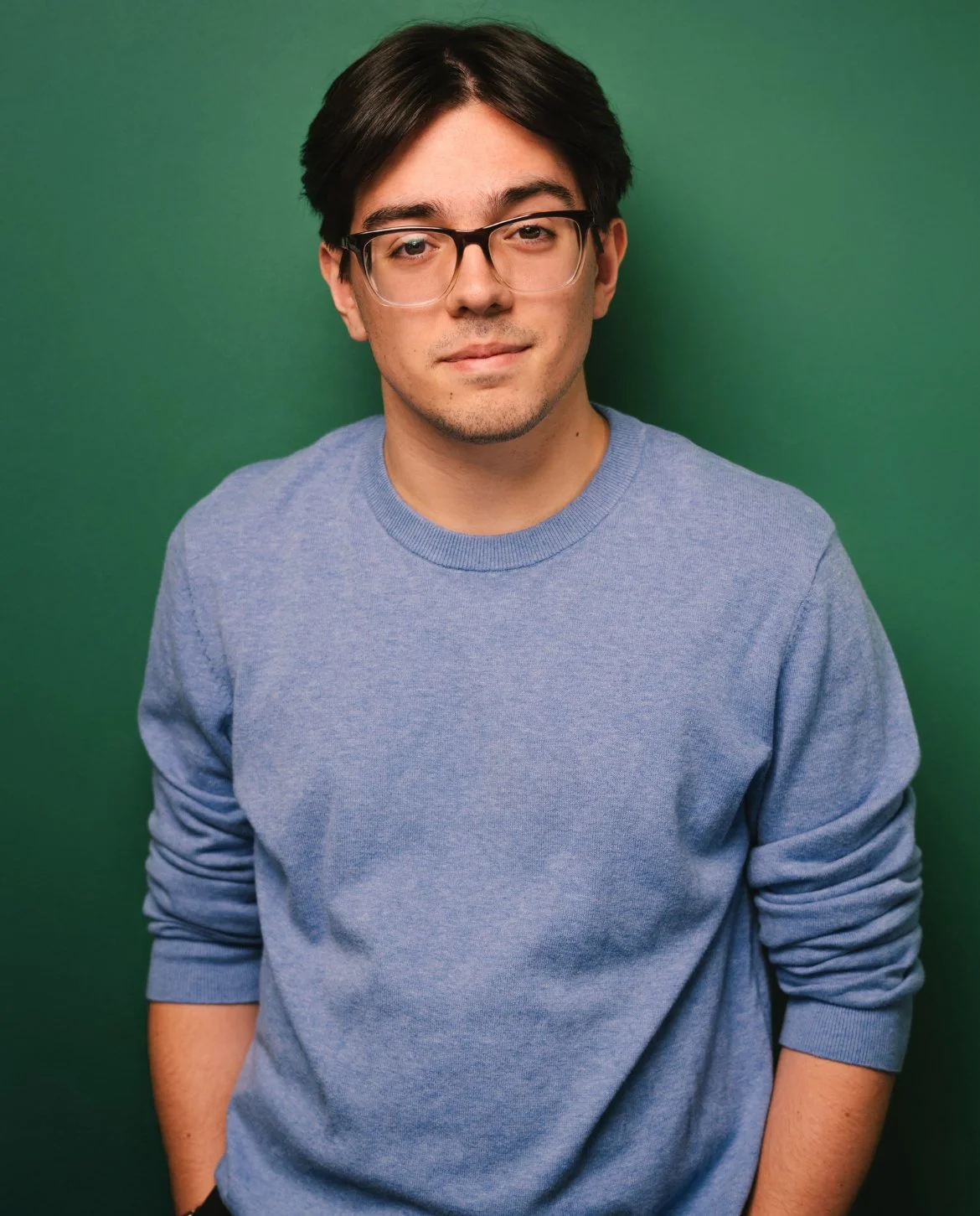 A young man with dark hair, glasses, and light skin, wearing a blue sweater, standing against a green background.