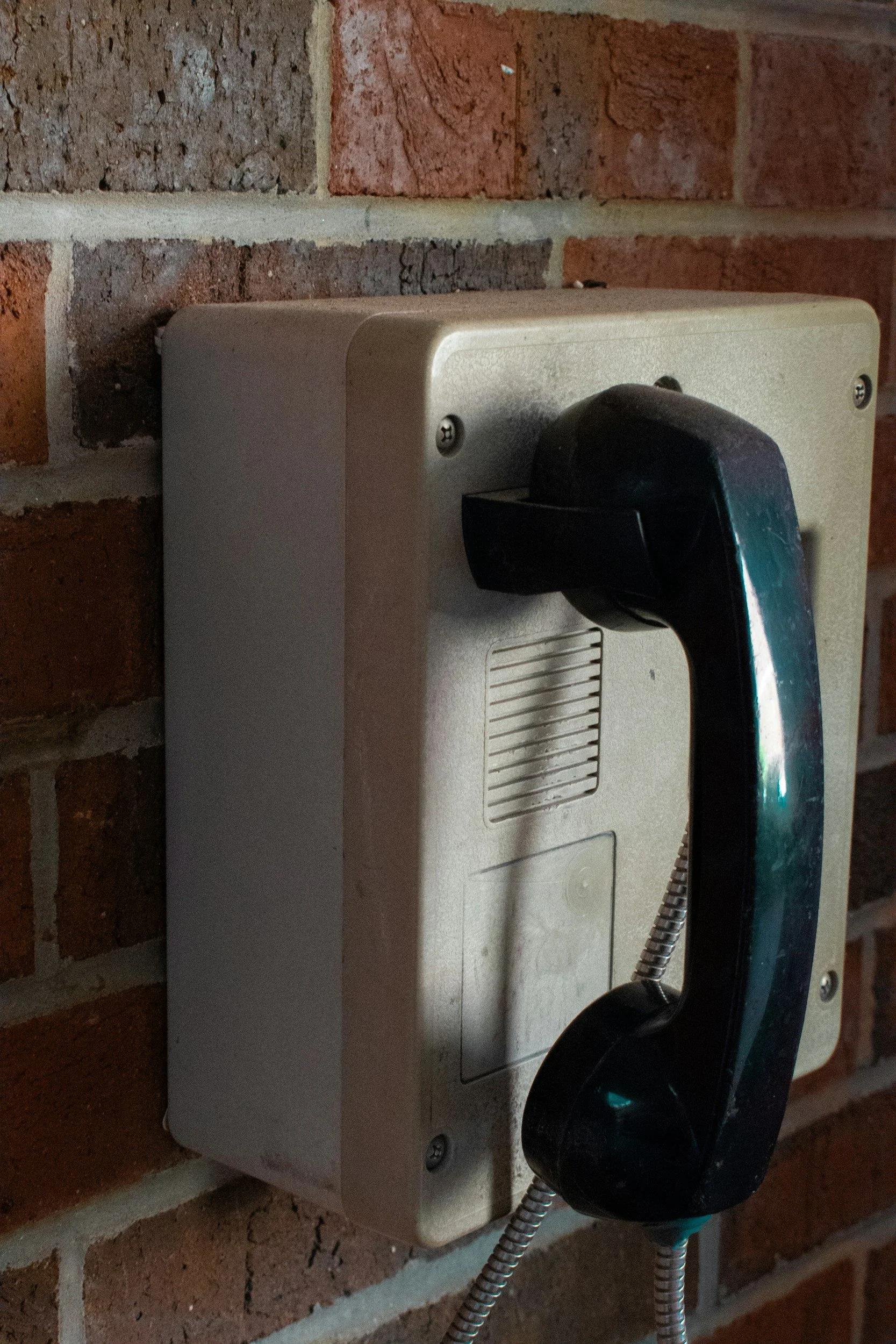 A beige wall-mounted telephone with a black receiver hanging on a metal cord, installed on a red brick wall.