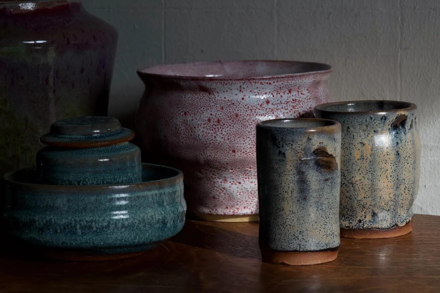Assorted ceramic pots and cups on a wooden surface against a wall, with natural light highlighting their textured glazes.