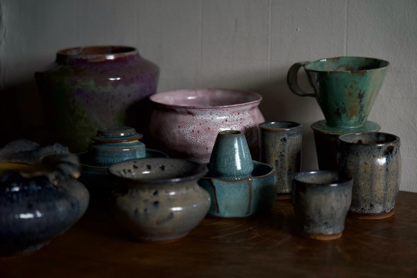 Various handmade ceramic vases and cups with muted earth tones and speckled glaze, arranged on a wooden surface against a plain wall.