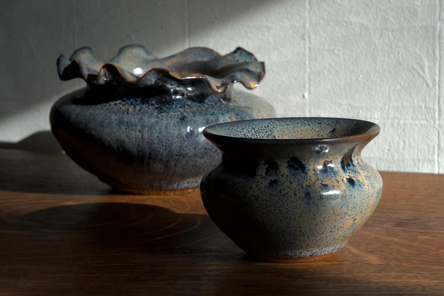 Two decorative ceramic vases with textured, speckled surfaces, one smaller and one larger, positioned on a wooden surface against a plain wall with shadows cast by the vases.