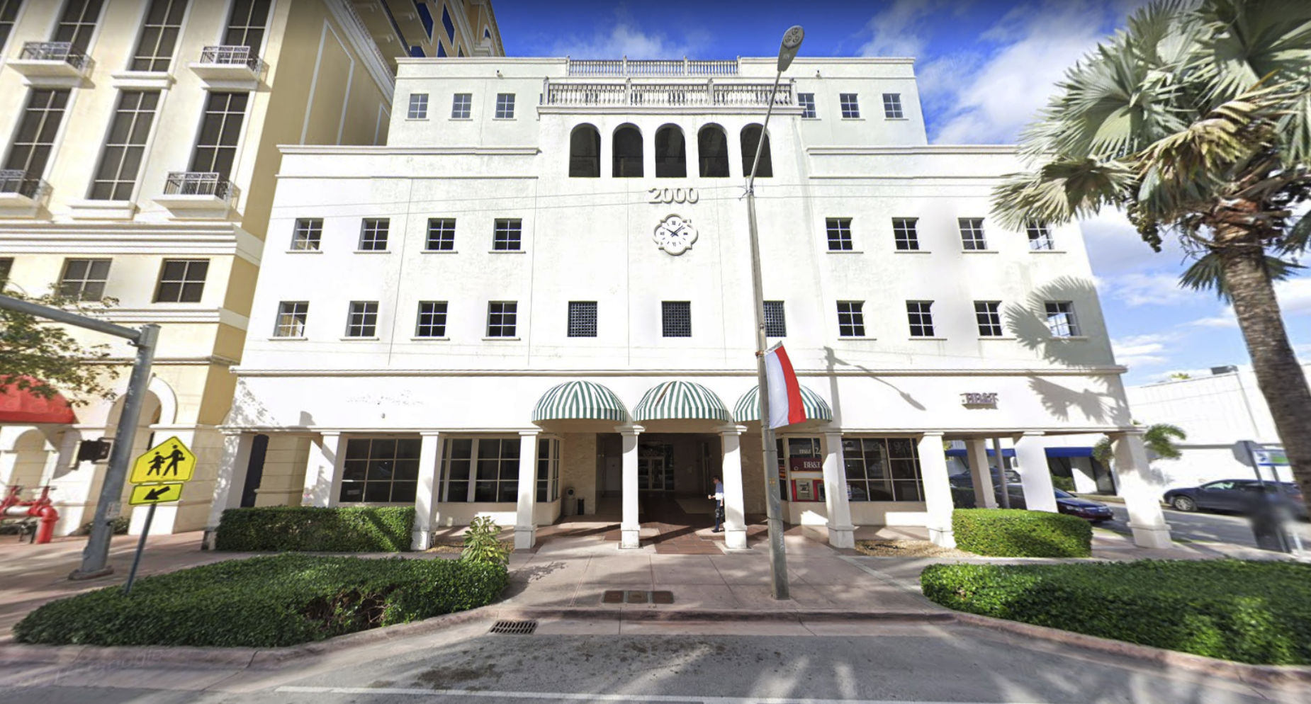 Front view of a white building with arched windows, a clock reading 11:19, green-striped awnings, and a tall palm tree on the right, under a partly cloudy sky.