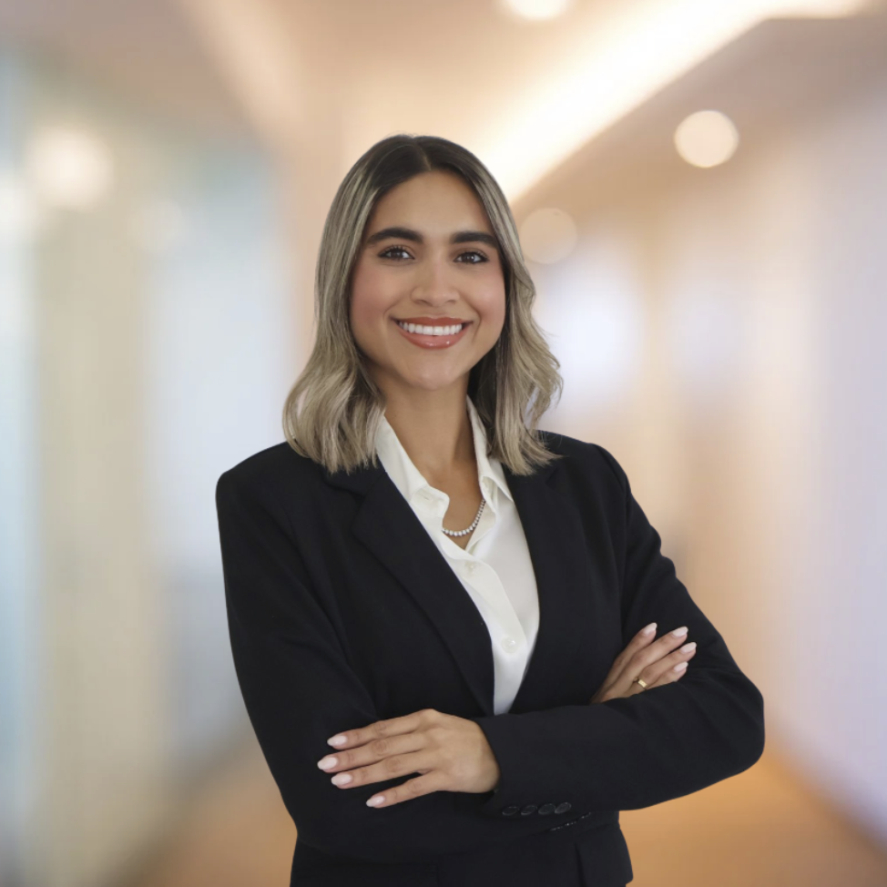 A woman with shoulder-length blonde hair, smiling, wearing a black blazer, white blouse, and pearl necklace, standing with arms crossed in a professional setting with blurred warm-colored background.