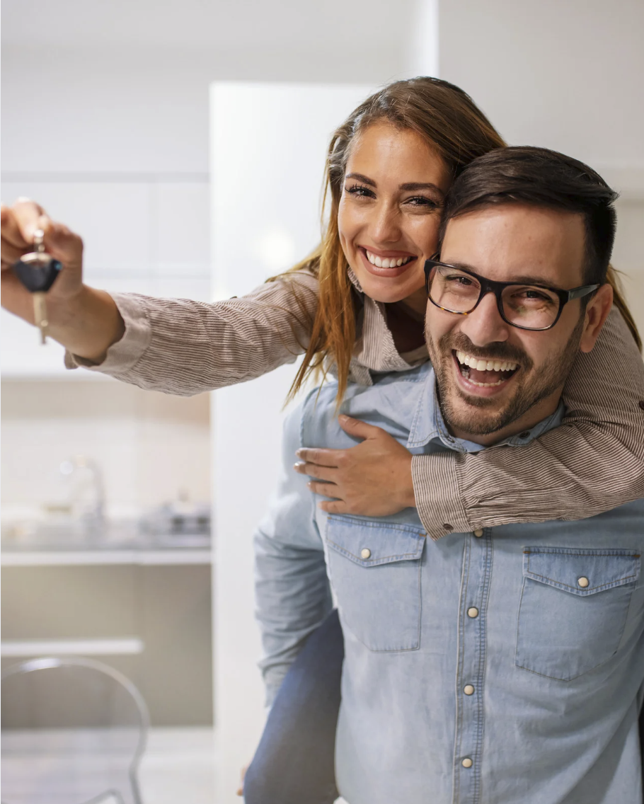 A smiling woman in a beige shirt giving a piggyback ride on a man's back while holding a set of keys, in a bright kitchen.