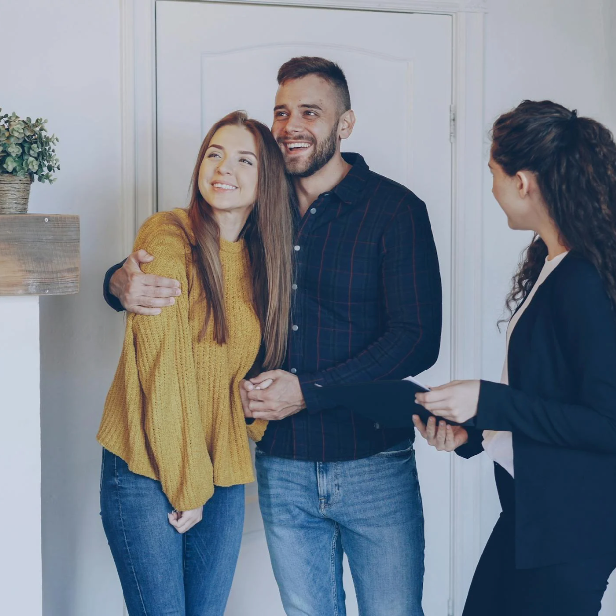 A couple hugging each other while a woman in business attire holds a clipboard and smiles, in a cozy home setting.