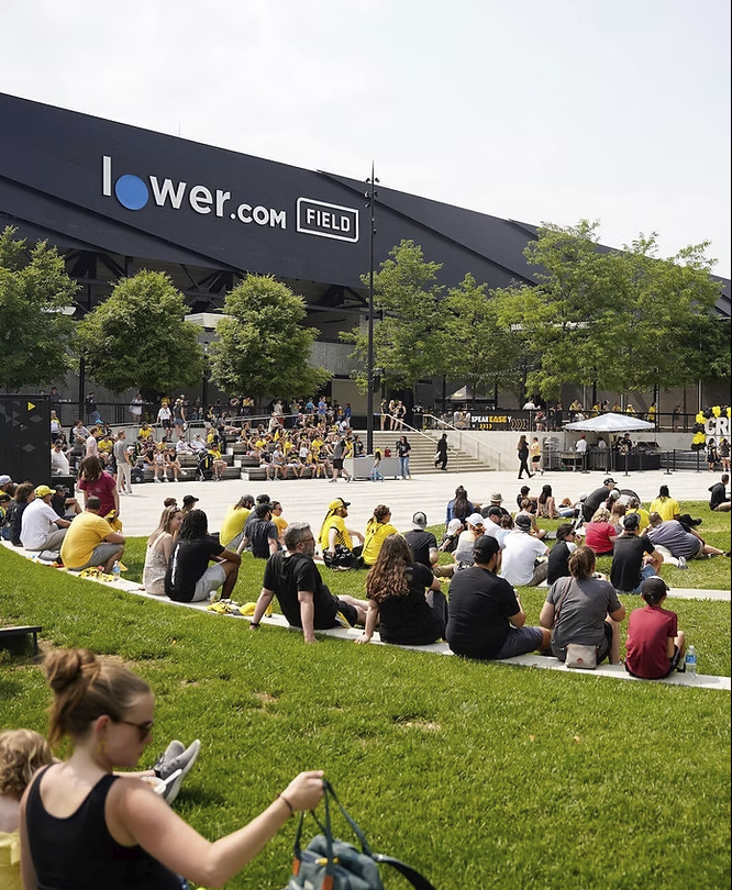 People sitting and standing on a grassy area and concrete steps outside a stadium, watching a performance or event.
