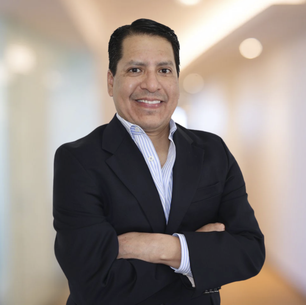 A man in a business suit with arms crossed, smiling, in front of a blurred indoor background.