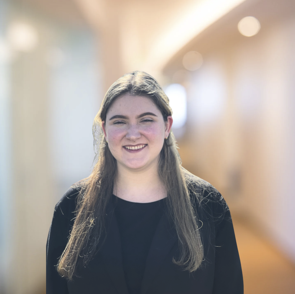 Young woman with long brown hair smiling, standing in a brightly lit corridor with soft-focus background.
