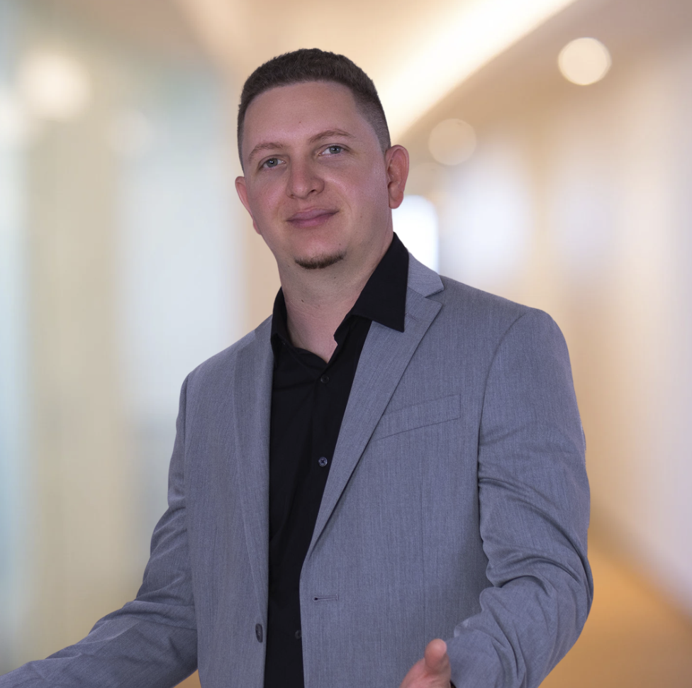 A young man with short hair, a goatee, wearing a gray blazer and black shirt, posing confidently in a professional setting with blurred warm-colored background.