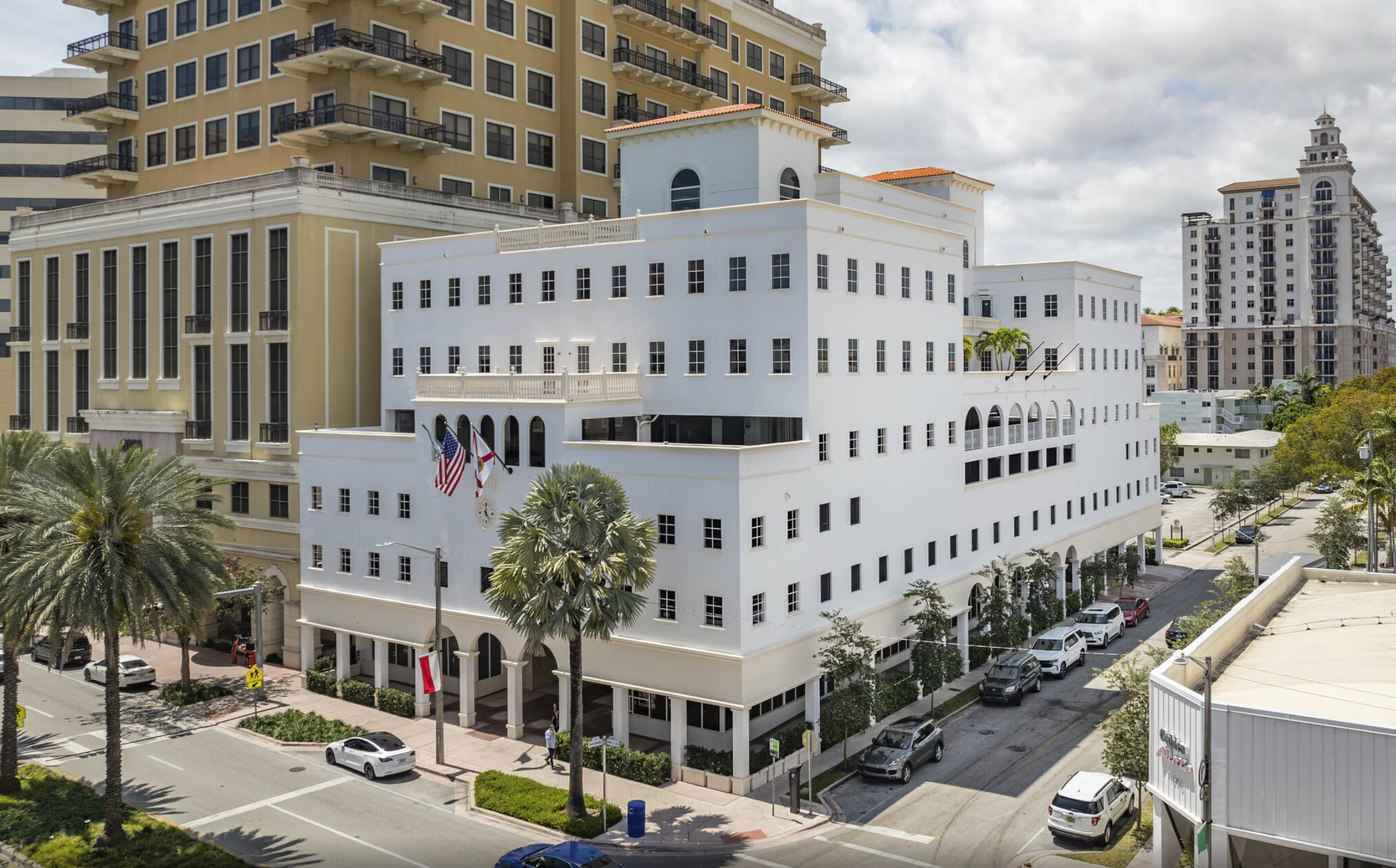 A white multi-story building with arched windows and palm trees in front, situated on a street with parked cars and surrounded by taller beige and brown buildings.