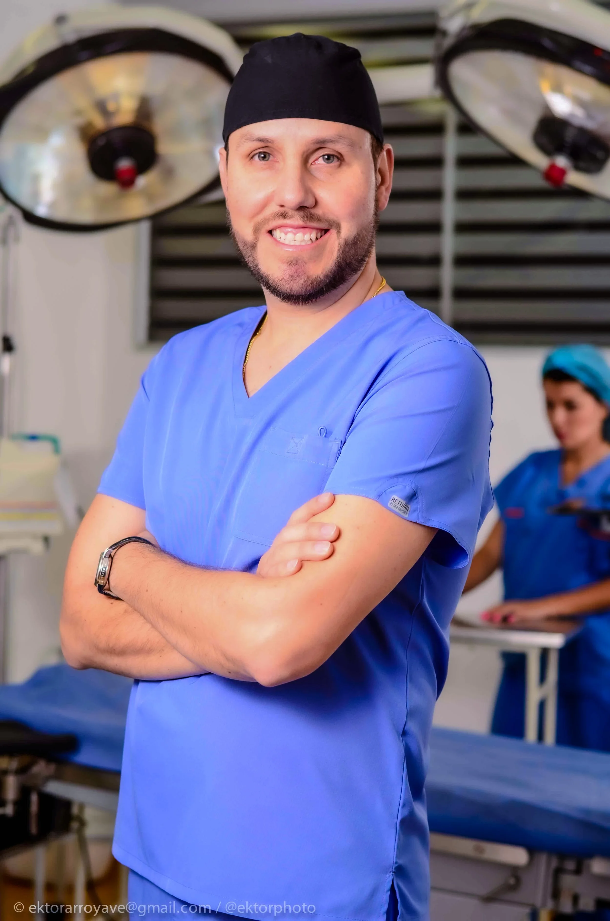 Un cirujano vestido con uniforme azul, gorro y con los brazos cruzados, en un quirófano, con luz quirúrgica y otro personal en el fondo.