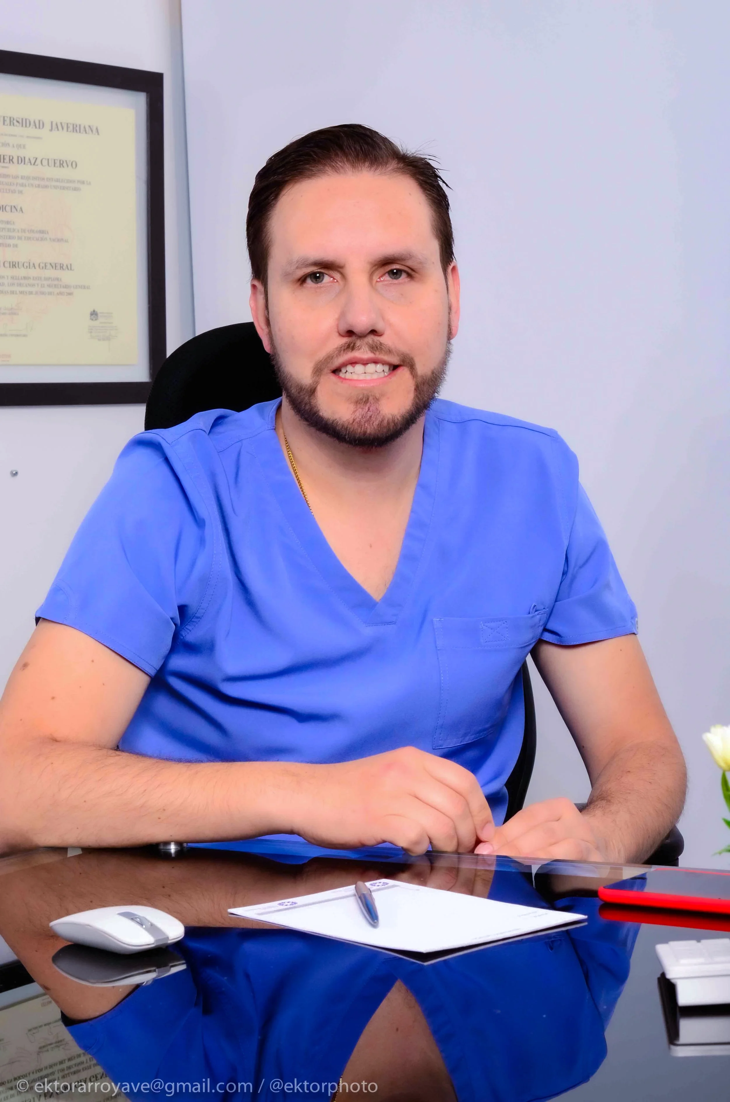 Un hombre con barba y cabello corto sentado en oficina, lleva puesta una bata azul y tiene una libreta y un bolígrafo sobre la mesa delante de él.